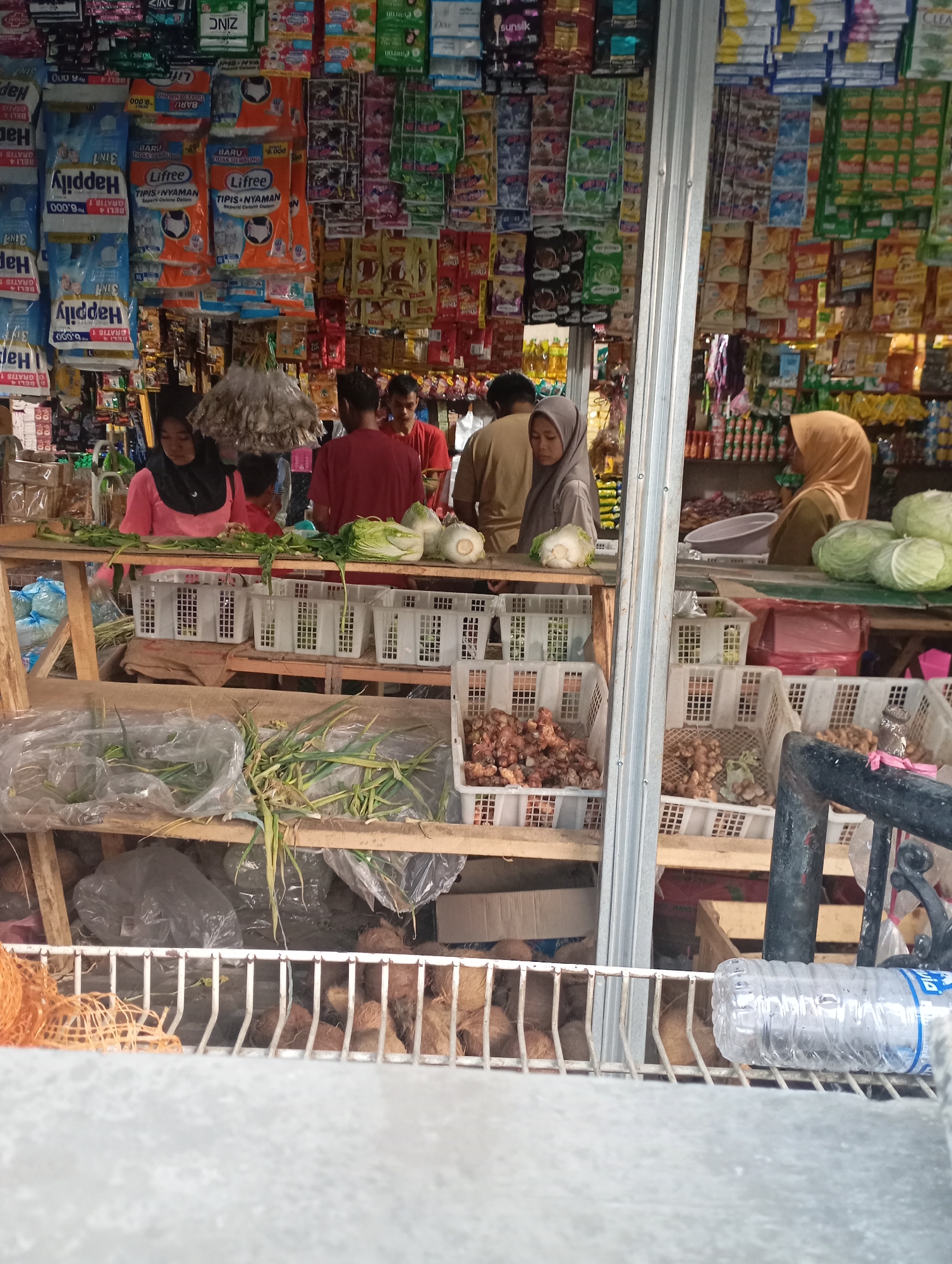 The image captures a street-side market stall in Kecamatan Haurgeulis, Indonesia. The stall is laden with various goods, primarily food items and packaged products. In the foreground, a display of coconuts is visible in a metal rack, with a plastic water bottle resting on the same rack. Above this, on wooden shelves, are baskets containing produce like green onions and what appear to be root vegetables or spices, possibly ginger. Further back, various packaged goods, including detergents, soaps, and food items in colorful wrappers, are prominently displayed on shelves and hanging from the stall's structure. Three individuals are present: a woman in a pink shirt and black headscarf is tending to the produce, another woman in a light brown hijab is standing near the produce, and a man in a red shirt is also present in the stall. The background suggests a bustling marketplace with more stalls and awnings, indicative of a typical Indonesian urban or semi-urban setting. The overall scene depicts a commercial transaction point within the local community.