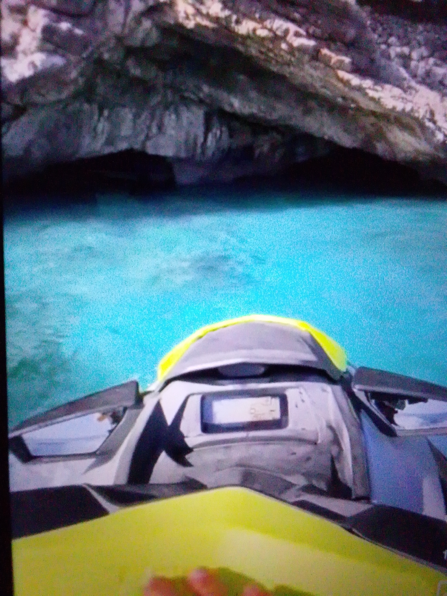 The image depicts a personal watercraft, commonly known as a jet ski, navigating through a body of water. The watercraft is primarily gray and black with a vibrant yellow accent on the front. It features a dashboard with a small display screen. The surrounding environment appears to be a natural setting, possibly a coastline or a waterway with rocky formations and cliffs overhead. The water is a bright turquoise color, indicating clarity and possibly shallow depth in some areas. The scene is viewed from the perspective of someone on the jet ski. The city of Aznakayevo, Russia, is not discernible from the visual elements of this image.
