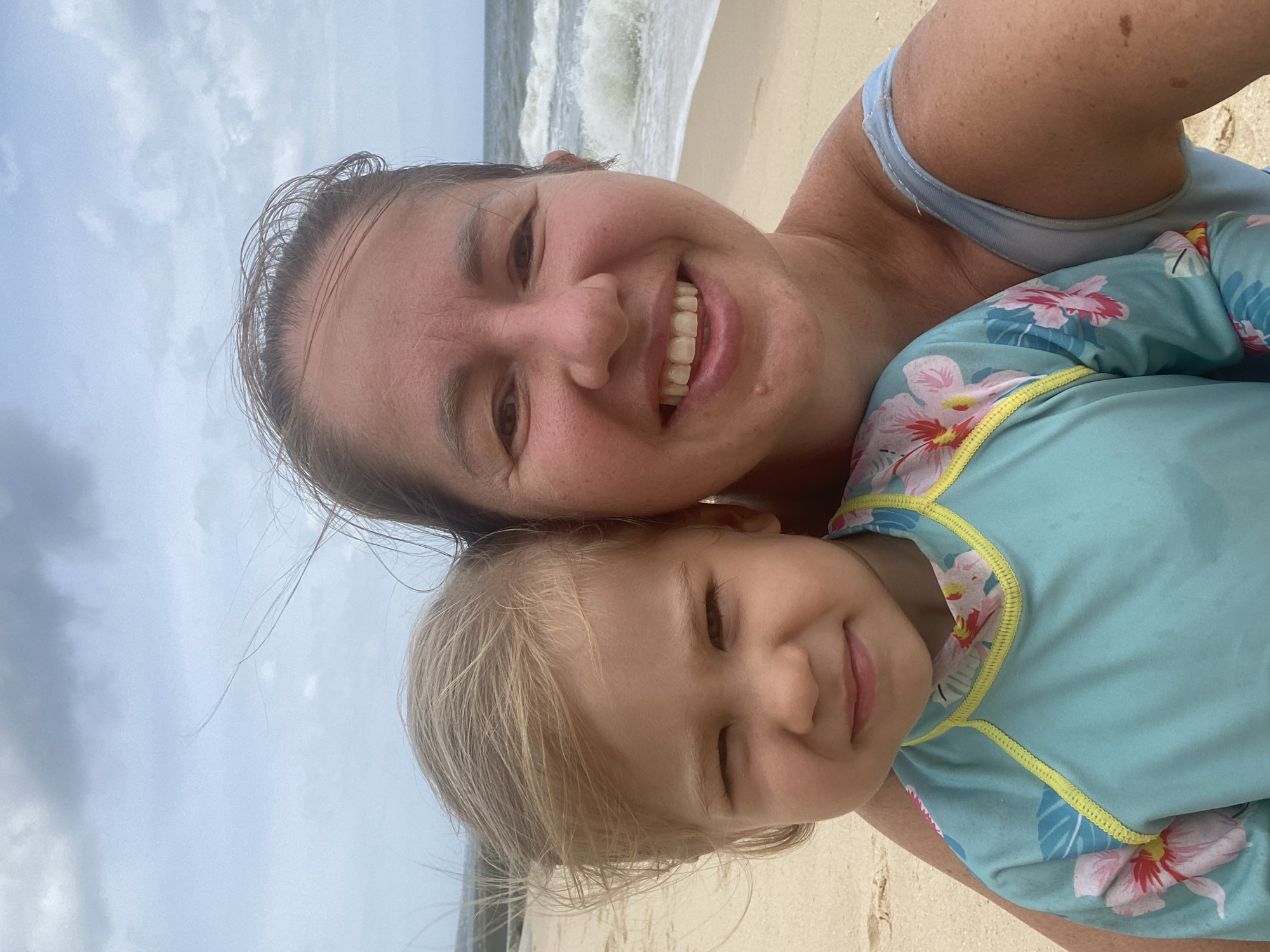 A close-up selfie shows a woman and a young child on a sandy beach. The woman is on the right, smiling broadly and looking at the camera. She has light brown hair and is wearing a blue swimsuit. The child is on the left, with blonde hair and a blue rash guard with a floral pattern. The child is looking at the camera with a slight squint. The background shows the ocean with waves and a cloudy sky. The location is identified as Mũi Né, Vietnam.