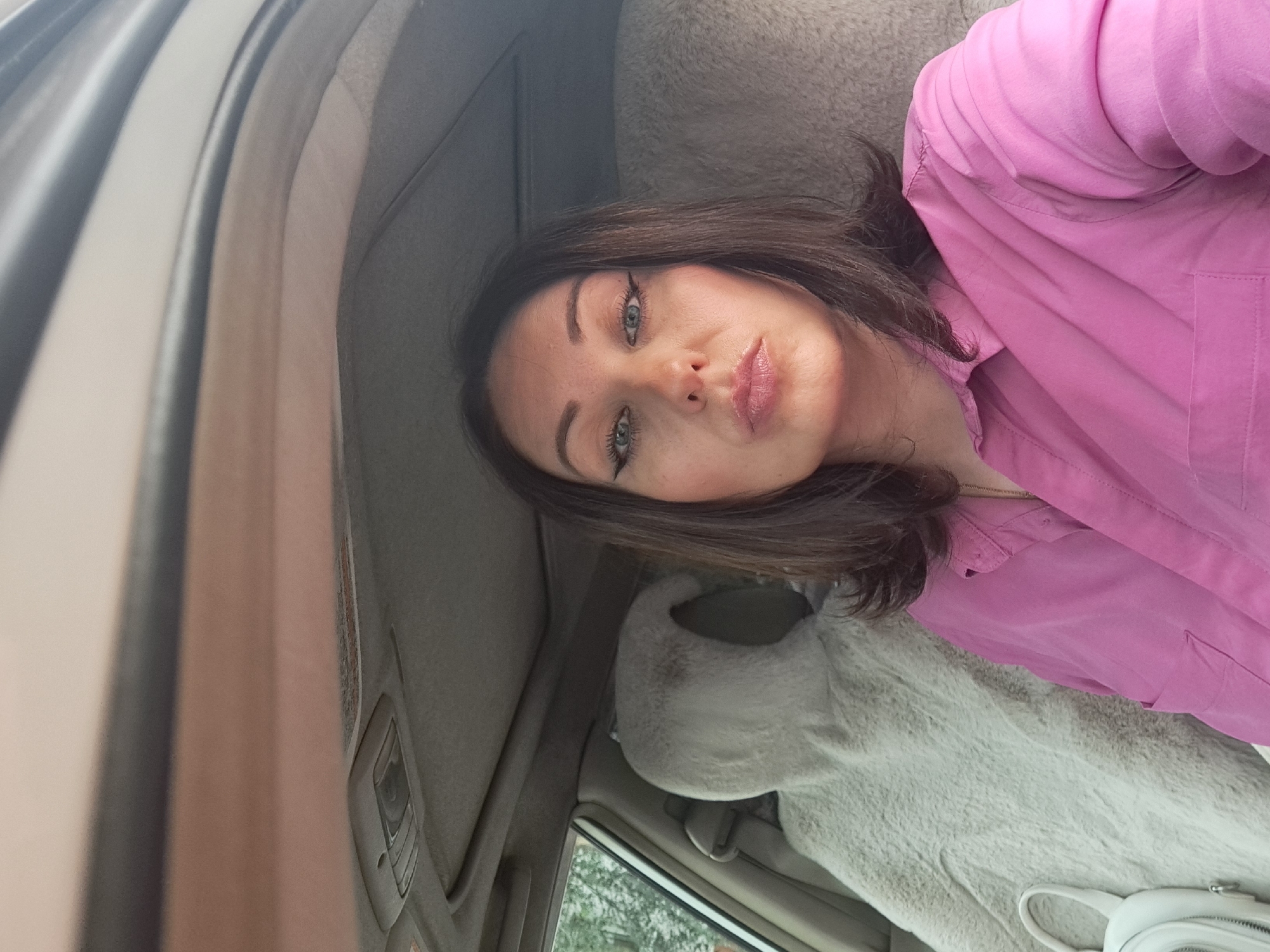 The image shows a woman in a car, taking a selfie. She is wearing a pink shirt and has dark brown hair. She is wearing makeup, including winged eyeliner and lip gloss. The car's interior is visible, with a fluffy grey seat cover on the passenger side. The background is blurred, but appears to be trees and foliage, suggesting an outdoor location. The image does not contain any specific landmarks or features that would allow for identification of the location within Irkutsk, Russia.