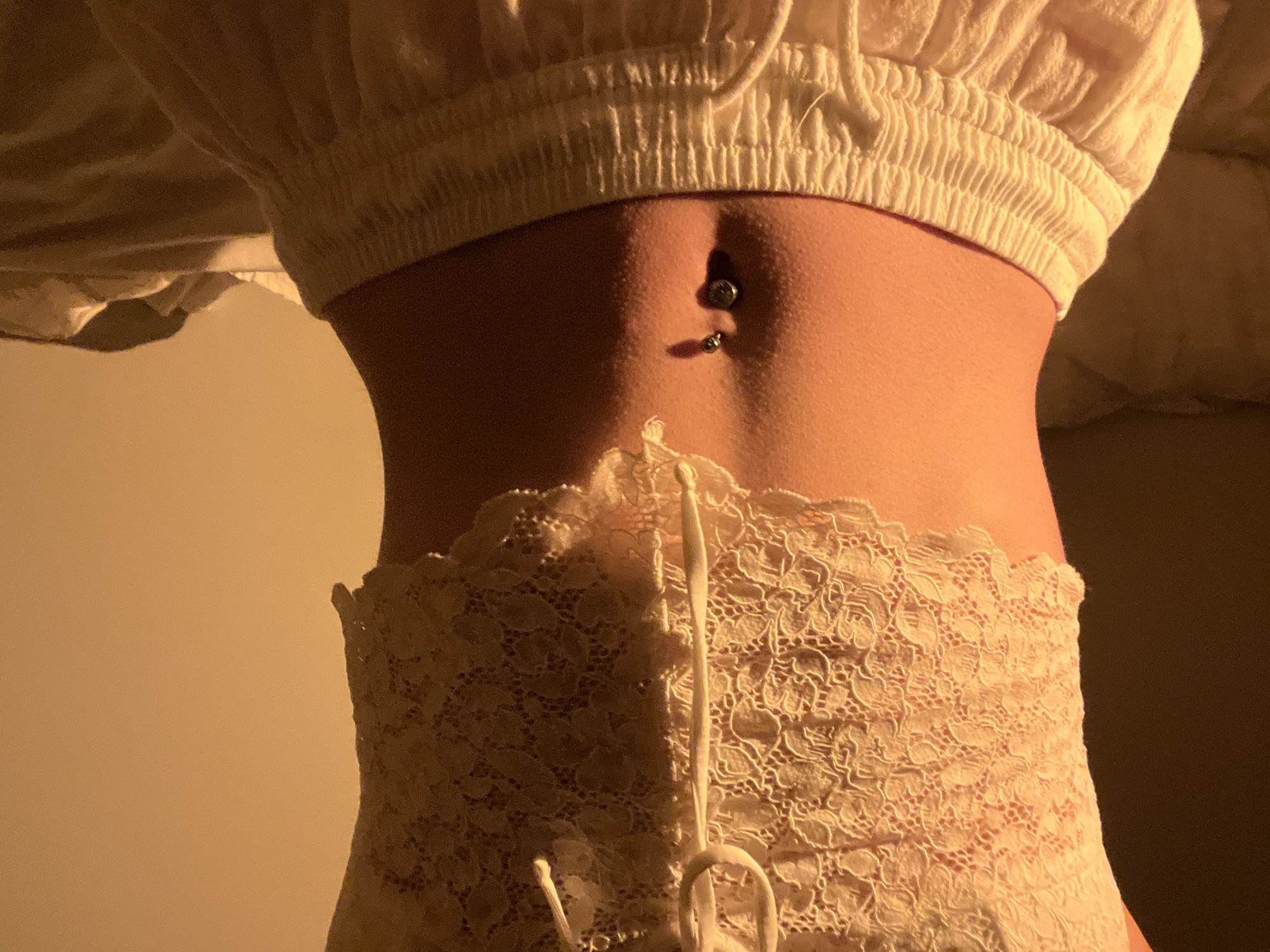 The image shows a close-up of a person's midriff, from just below the bust to the upper thighs. They are wearing a light-colored, lacy, cropped top with a drawstring at the front, and loose-fitting, light-colored sweatpants with a drawstring. The person has a navel piercing with a silver belly ring. The lighting is warm and golden, creating shadows that highlight the contours of the abdomen. The background is indistinct and appears to be a bed with rumpled sheets. There is no indication of the city of Ternopil, Ukraine, or any urban environment.