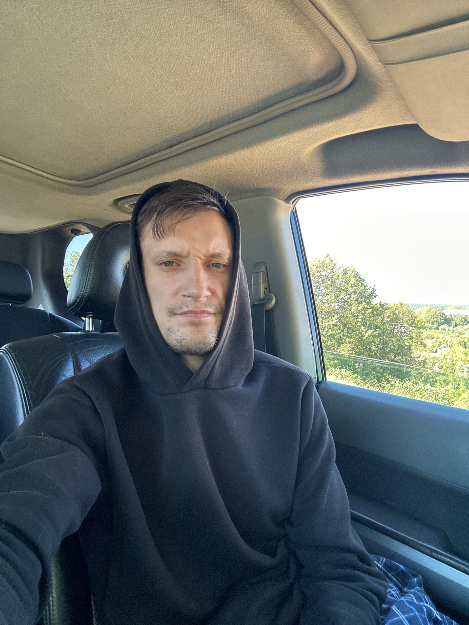 A man in a black hoodie takes a selfie from the driver's seat of a car. He has short brown hair and stubble on his chin. The car has a sunroof open, and through the open window, a scenic landscape of green trees and rolling hills is visible. The interior of the car is dark, with black leather seats and a beige headliner. The man is looking directly at the camera with a neutral expression. The image appears to have been taken on a sunny day. There is no information about the location in the city of Makiv, Ukraine.