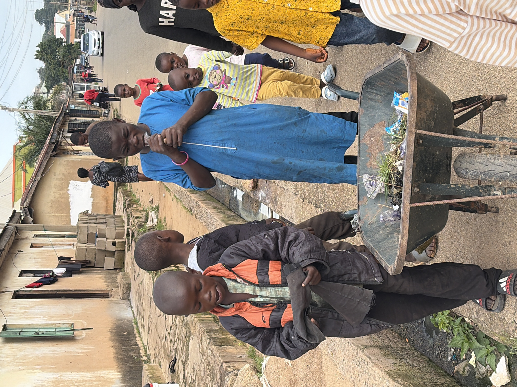 In Jos, Nigeria, a group of children are gathered on a street. A young boy in a blue robe is holding a piece of material to his mouth. To his left, another boy in a black and orange jacket is holding a dark cloth. In the foreground, a wheelbarrow filled with debris sits on the ground. Other children, dressed in various casual clothing, are visible in the background, some walking along the street. The scene appears to be in a residential area with modest buildings and a paved road.