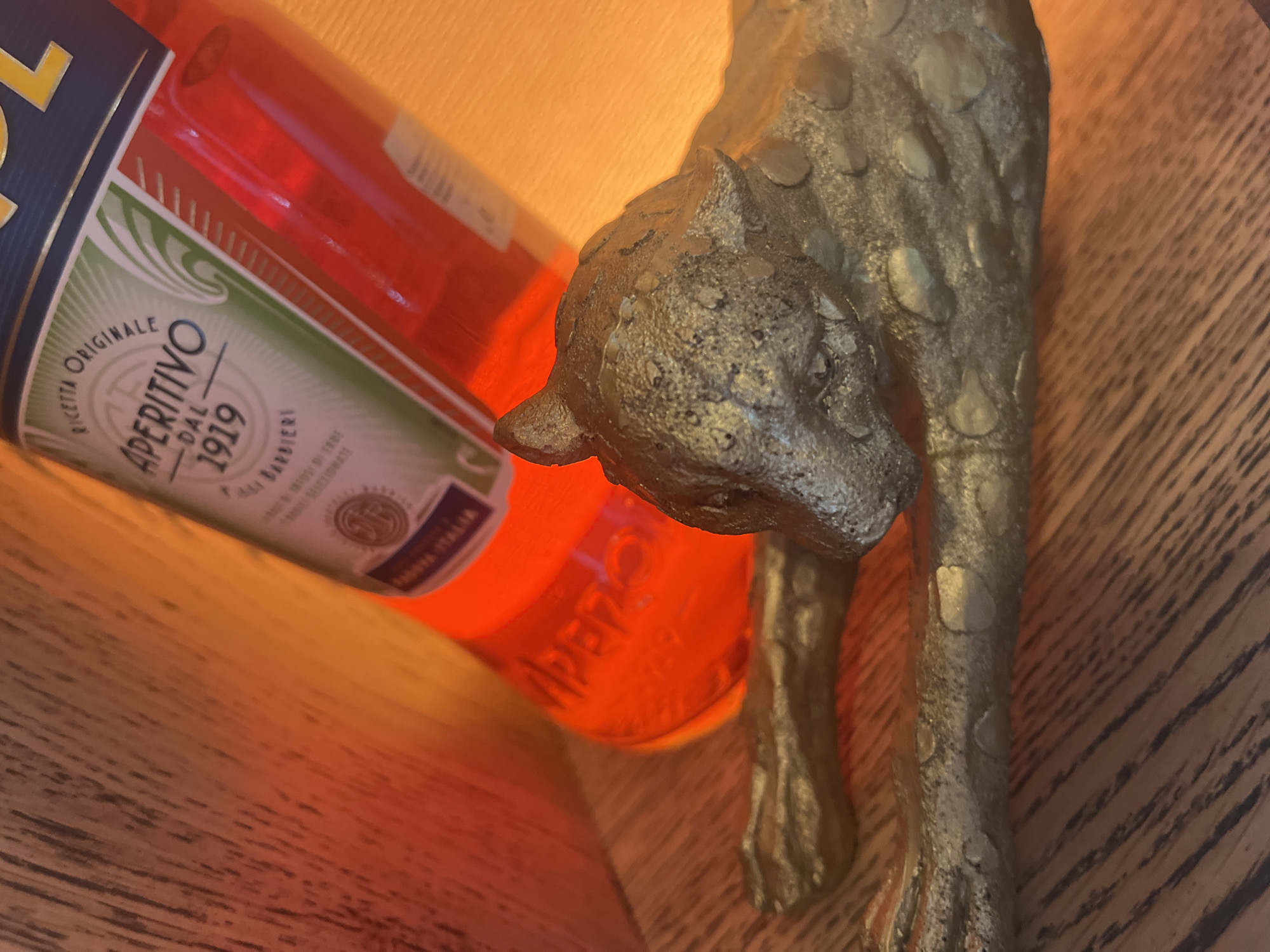 The image features an angled, close-up view of a vibrant, translucent orange-red bottle, identifiable as Aperitivo, positioned next to a decorative metallic animal figurine. The bottle's label is prominently displayed, showcasing text including "RICETTA ORIGINALE", "APERITIVO", "DAL 1919", and "BARBIERI", along with "PRODOTTO IN ITALIA". The word "APERO" is also visibly embossed into the glass of the bottle near its base.
To the right, partially in the foreground, is a gold-toned figurine of an animal, likely a cheetah or leopard, with a rough, textured, and spotted surface. Its head is angled downwards, and its body extends into the frame, adorned with raised, irregular metallic spots. The figure has an antique or handcrafted appearance.
The scene is set indoors, on a wooden surface with a visible grain pattern, possibly a table or shelf. The background is softly illuminated by a warm, yellowish-orange light, creating an inviting and cozy ambiance. The overall impression is that of a decorative still life, with no specific activity taking place. The warm lighting suggests an evening or artificial indoor setting.