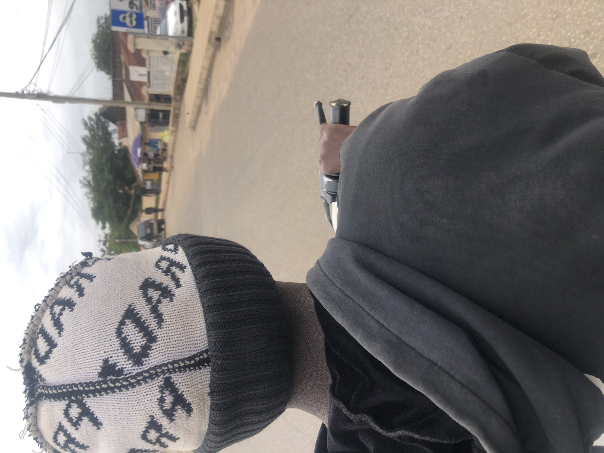 A person wearing a black and white patterned beanie and a grey hoodie is riding a motorcycle on a paved road. The person is holding the motorcycle's handlebars with their right hand. In the background, there are buildings, trees, and a cloudy sky. Some people and vehicles are visible in the distance. The overall scene appears to be a street in a city or town.