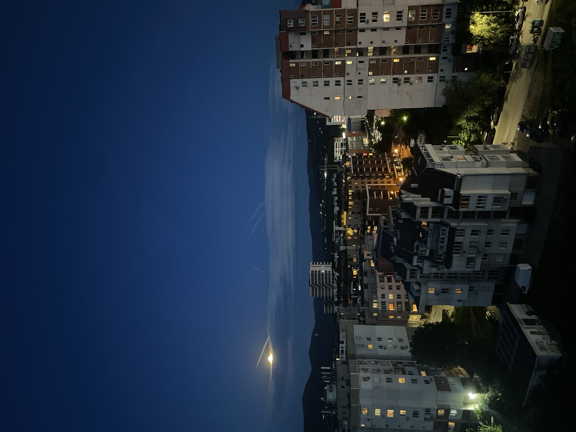 This image depicts a nighttime urban scene in Sofia, Bulgaria. The view is oriented vertically, capturing a large expanse of dark blue sky on the left, transitioning to a strip of land with hills and a cityscape on the right. The moon is visible near the horizon, with faint contrails in the sky above it. The cityscape is composed of several buildings of varying heights, predominantly apartment complexes, illuminated by interior lights. Streetlights are present, illuminating the roads and adjacent areas. Vehicles are parked along the streets. The architecture is typical of post-communist urban development in Eastern Europe.