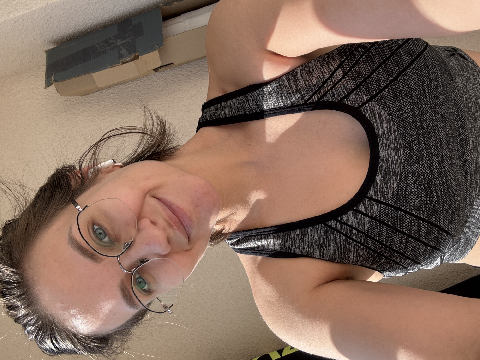 The image captures a selfie of a young woman with light brown hair and green eyes, looking directly at the camera with a subtle, closed-mouth smile. She wears wire-rimmed glasses and a white earbud is visible in her left ear. She is dressed in a grey and black patterned sports bra or athletic top, featuring a speckled texture with diagonal black striped sections.
Bright, natural sunlight illuminates the scene, casting distinct shadows on her skin and clothing, indicating a sunny daytime setting, likely morning or afternoon. The background consists of a light-colored, textured surface, possibly a carpet or rug. In the upper left corner, a worn, partially open cardboard box secured with blue tape is visible on this surface.
The overall impression is one of a casual, relaxed moment indoors in Pervomaysky, Russia. No specific activity is taking place other than posing for the selfie. There is no visible text in the image.