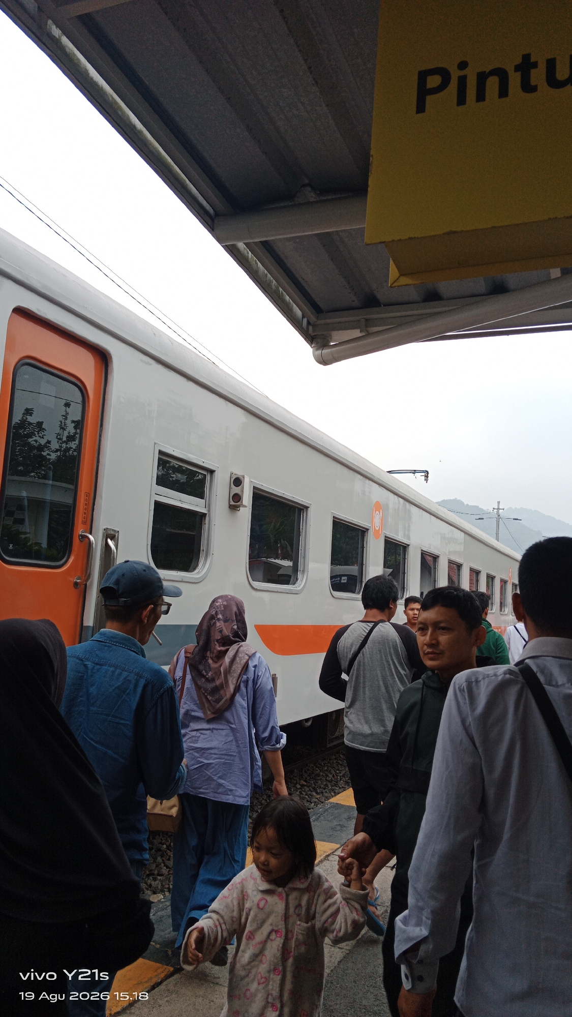 The image captures a scene at a train station platform in Garut, Indonesia. A passenger train, predominantly white with orange accents on its doors and along its lower body, is stopped at the platform. Several people are visible, appearing to be passengers. In the foreground, a young girl in a patterned fleece outfit is being held by the hand by an adult. Other individuals, including men and women, are standing and walking on the platform, some facing the train, others in profile. The platform is paved with yellow and grey tiles. Above, a grey canopy supported by metal structures shelters the platform. A yellow sign with black text, partially visible and reading "Pintu", is mounted on the canopy. The background suggests an overcast sky and possibly distant hills.
