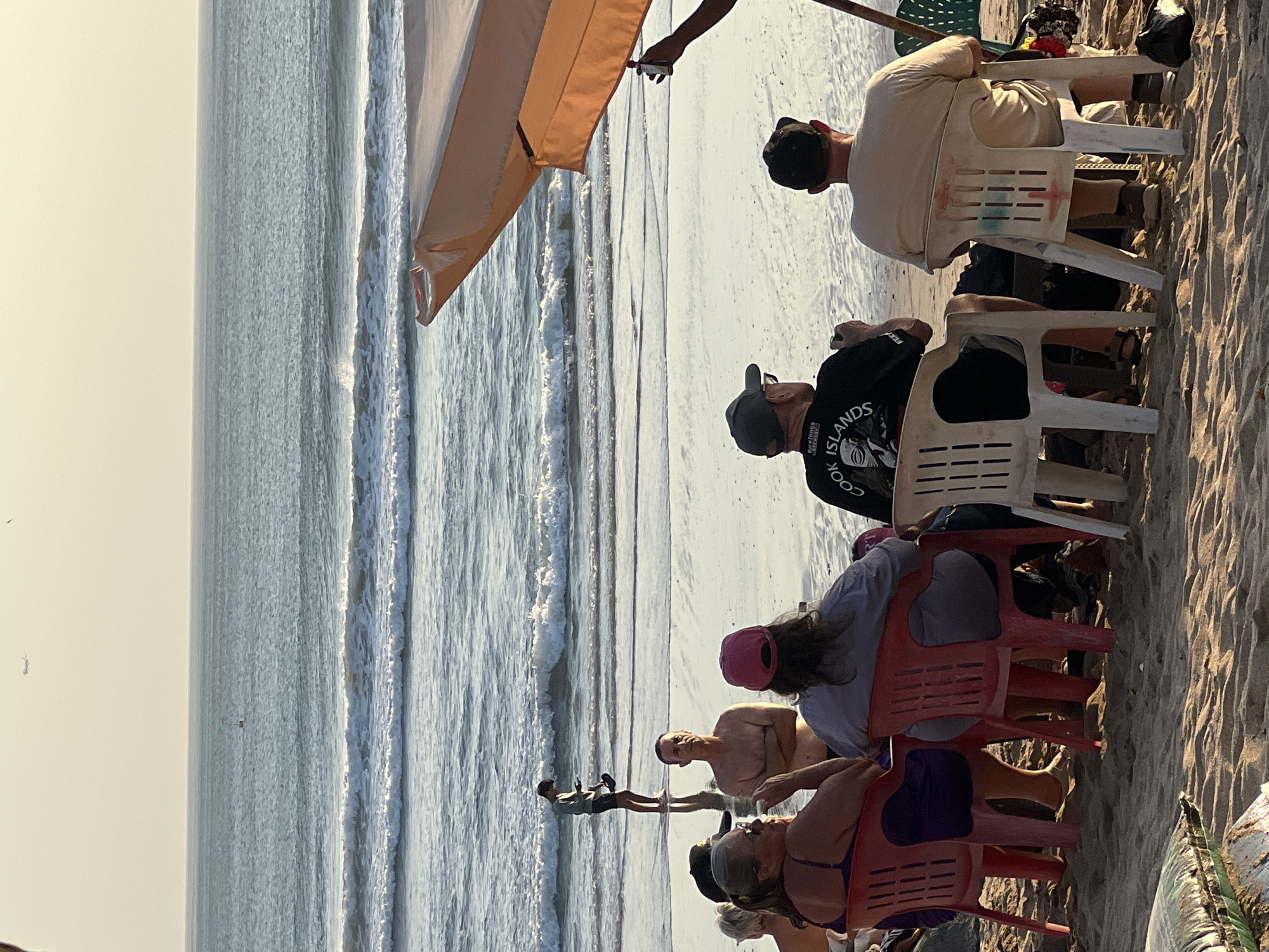 The image depicts a group of people on a beach in Kuta, Indonesia, likely during sunset. The foreground features several individuals seated on plastic chairs facing the ocean. Some are engaged in conversation, while others are looking out at the water. One person is standing in the shallow water, holding a phone. In the background, waves are breaking on the shore under a hazy sky. An orange beach umbrella is partially visible on the right side of the frame. The scene suggests a relaxed and casual gathering on a popular tourist beach.