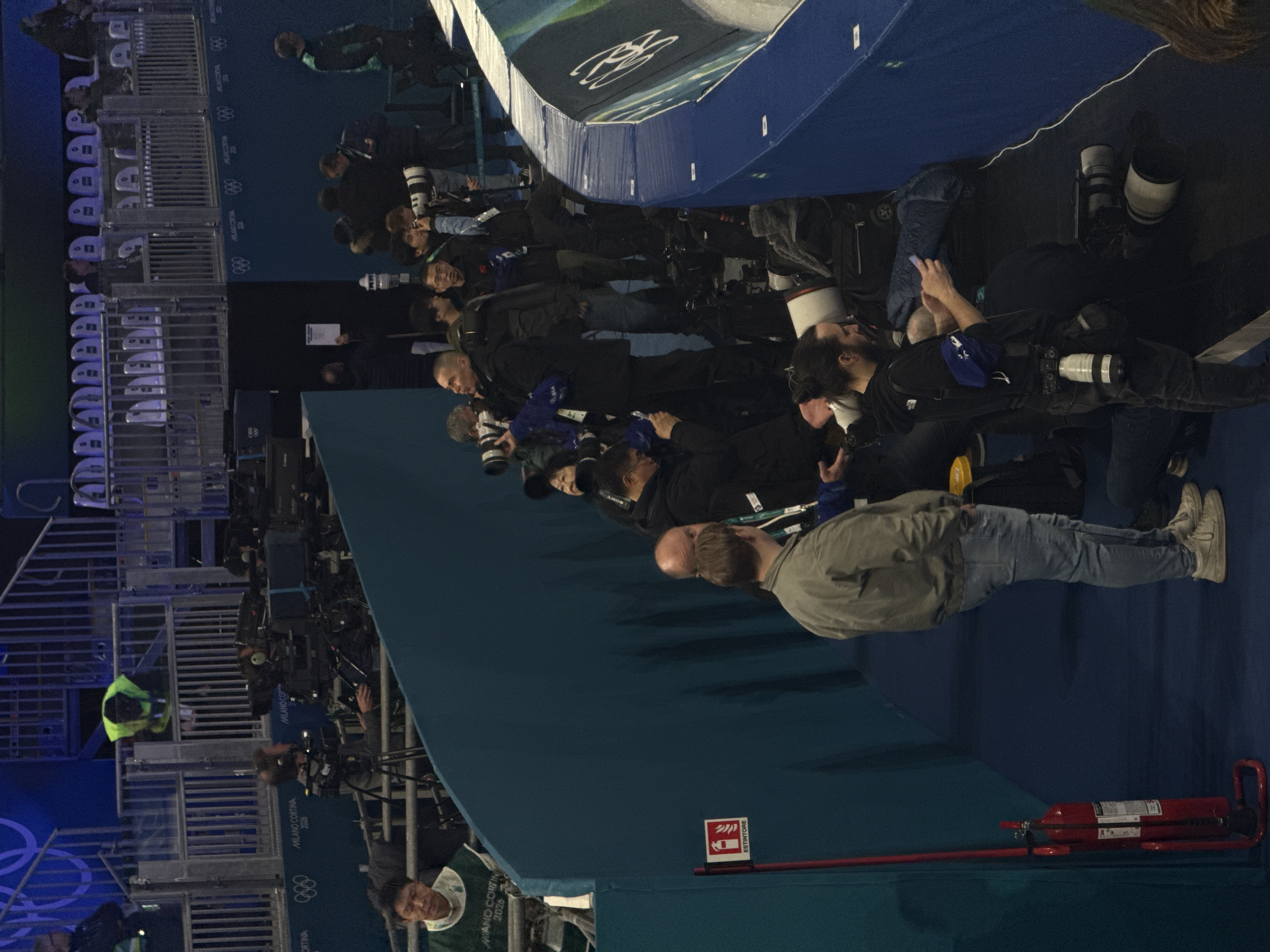 The image displays an indoor media zone within an event venue in Rho, Italy, associated with the Milano Cortina 2026 Winter Olympics. Approximately twenty to thirty individuals, predominantly adult males, are present. Many are professional photographers and videographers, equipped with various digital cameras fitted with large telephoto lenses, and several video cameras mounted on tripods. Individuals are positioned along barriers, either standing or kneeling, with their equipment oriented towards an unseen central event area. Some wear identification lanyards, and one person wears a green vest branded "MILANO CORTINA 2026."
The immediate foreground features a dark teal barrier, while a padded light blue and white barrier bearing the "MILANO CORTINA 2026" logo and Olympic rings runs along the right side. In the background, tiered spectator seating with light-colored chairs is visible behind metal railings. A prominent red "ESTINTORE" (fire extinguisher) sign is mounted on the left, next to a red fire extinguisher on a stand. The floor throughout the media area is a dark blue surface. The scene is illuminated by artificial indoor lighting.