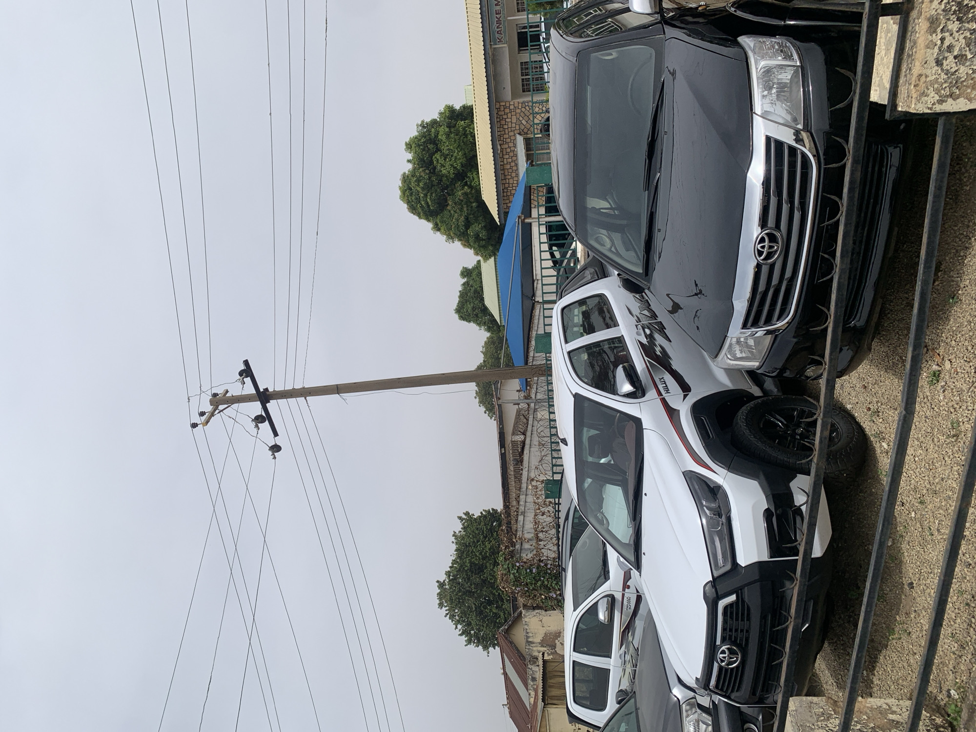 The image shows a row of parked vehicles, primarily Toyota Hilux trucks, in what appears to be a commercial area in Jos, Nigeria. The trucks are a mix of black and white models. An overhead power pole with numerous electrical wires is visible in the background, extending into a cloudy sky. Behind the vehicles, a building is seen with the name "KANKEME" visible on its facade. A fence runs parallel to the vehicles, partially obstructing the view. The environment suggests a vehicle dealership or a commercial parking lot.