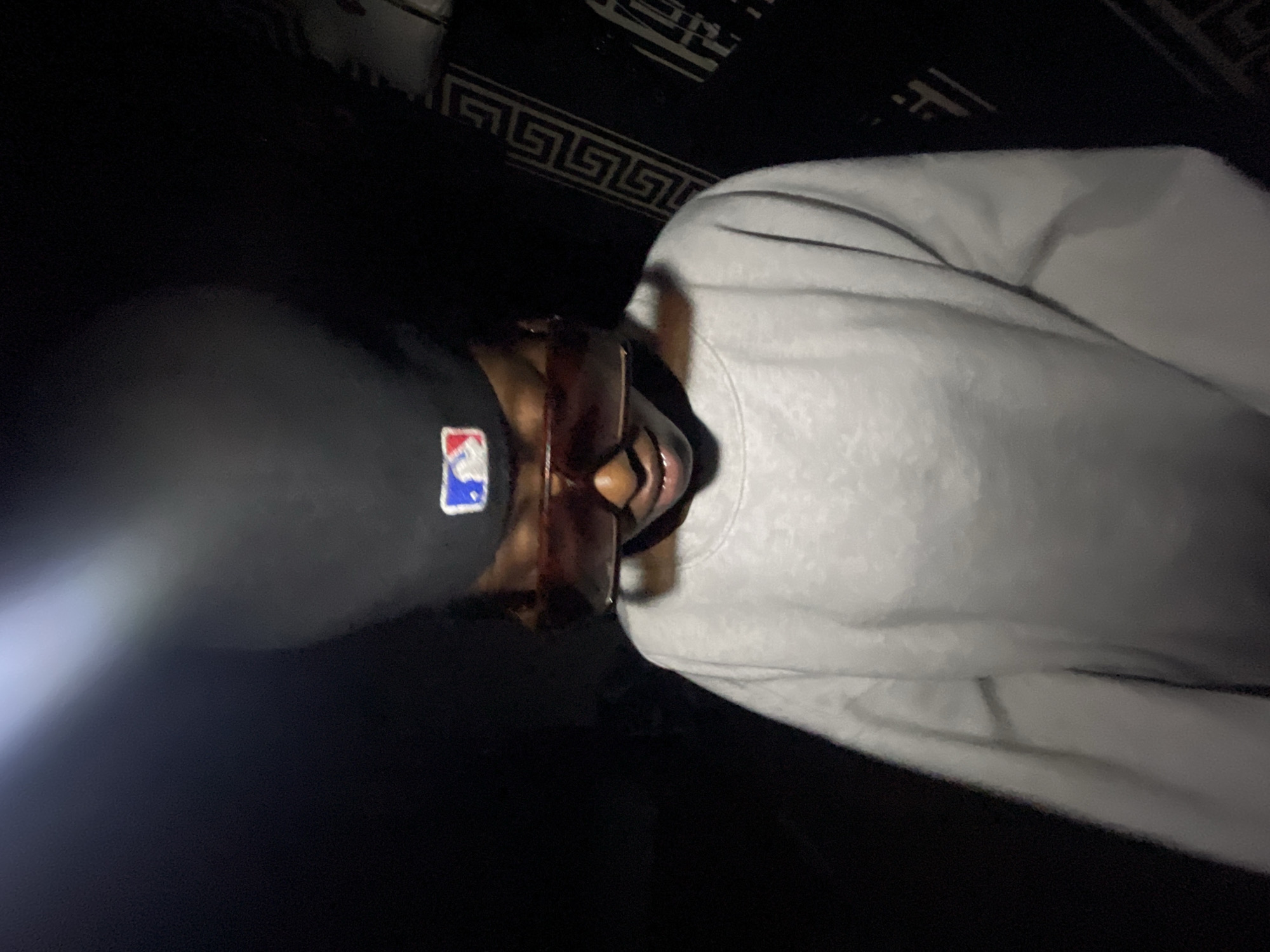 The image shows a close-up of a person wearing a black baseball cap with a red, white, and blue logo and oversized tinted sunglasses. They are also wearing a light-colored, long-sleeved sweatshirt. The background is dark, with a patterned fabric on the right side. The lighting suggests the image was taken in a dimly lit environment, possibly at night, with a bright light source originating from the upper left, creating a lens flare effect. The person's head is slightly bowed, and their facial features are obscured by the sunglasses and the angle of the shot. There is no discernible information in the image that would allow for the identification of a specific location in Lokoja, Nigeria.