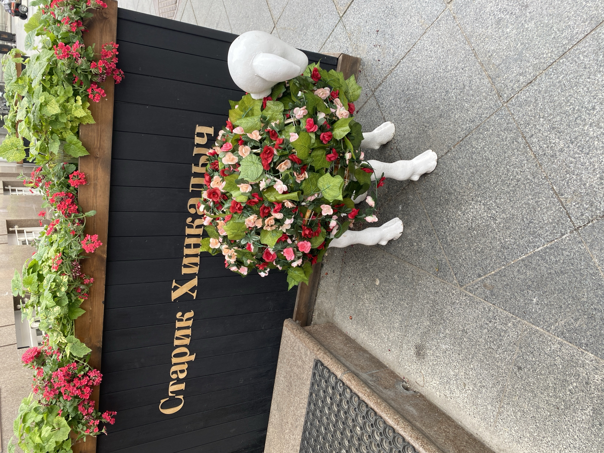 The image shows a street-level view of a decorative display in Moscow, Russia, likely near the entrance of a restaurant. The display consists of a black wooden structure with vertical slats, adorned with the Cyrillic text "Старик Хинкалыч" written in gold lettering. A window box overflowing with green plants and red flowers is attached to the left side.
To the right of the sign is an unusual sculpture of a person, mostly obscured by a large floral arrangement composed of red and pink roses and greenery. Only the white-painted head and limbs of the figure are visible. The sculpture is positioned on the gray, tiled sidewalk. A black rubber mat is visible in the lower right corner.