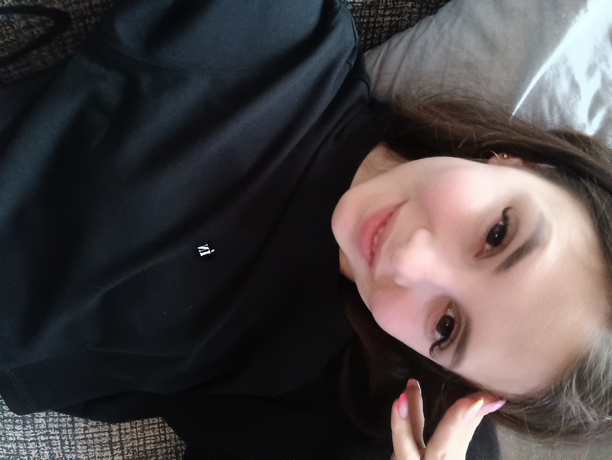 A woman is lying down, facing the camera, wearing a black long-sleeved shirt with a small, white, inverted logo on the upper left. Her hair is dark brown, and she has light skin and dark eyes. She is partially resting on a grey pillow or cushion. The background appears to be a textured grey surface, possibly a couch. The location is not discernible from the image, therefore, it is impossible to confirm if it's Chaikovsky, Russia.