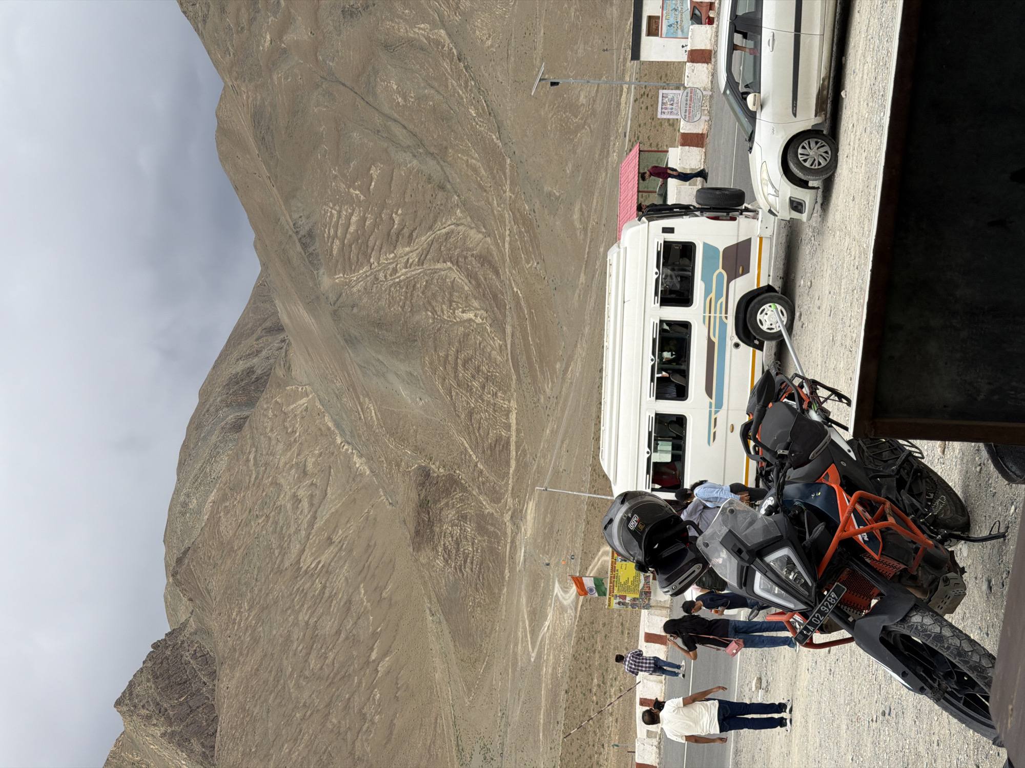 In Chumkar, India, a mountainous landscape dominates the background under a cloudy sky. In the foreground, a black and orange KTM motorcycle is parked on a gravel surface. A white van and a white car are positioned to the right of the motorcycle. Several people are visible: two men and a woman stand near the white van, a man in a blue checkered shirt walks away from the camera on the left, and another man in a white t-shirt and jeans walks towards the camera on the left. A woman in a blue shirt and jeans stands near the motorcycle. A small Indian flag is displayed on a pole near a sign. The overall scene depicts a roadside stop in a scenic, arid mountainous region.