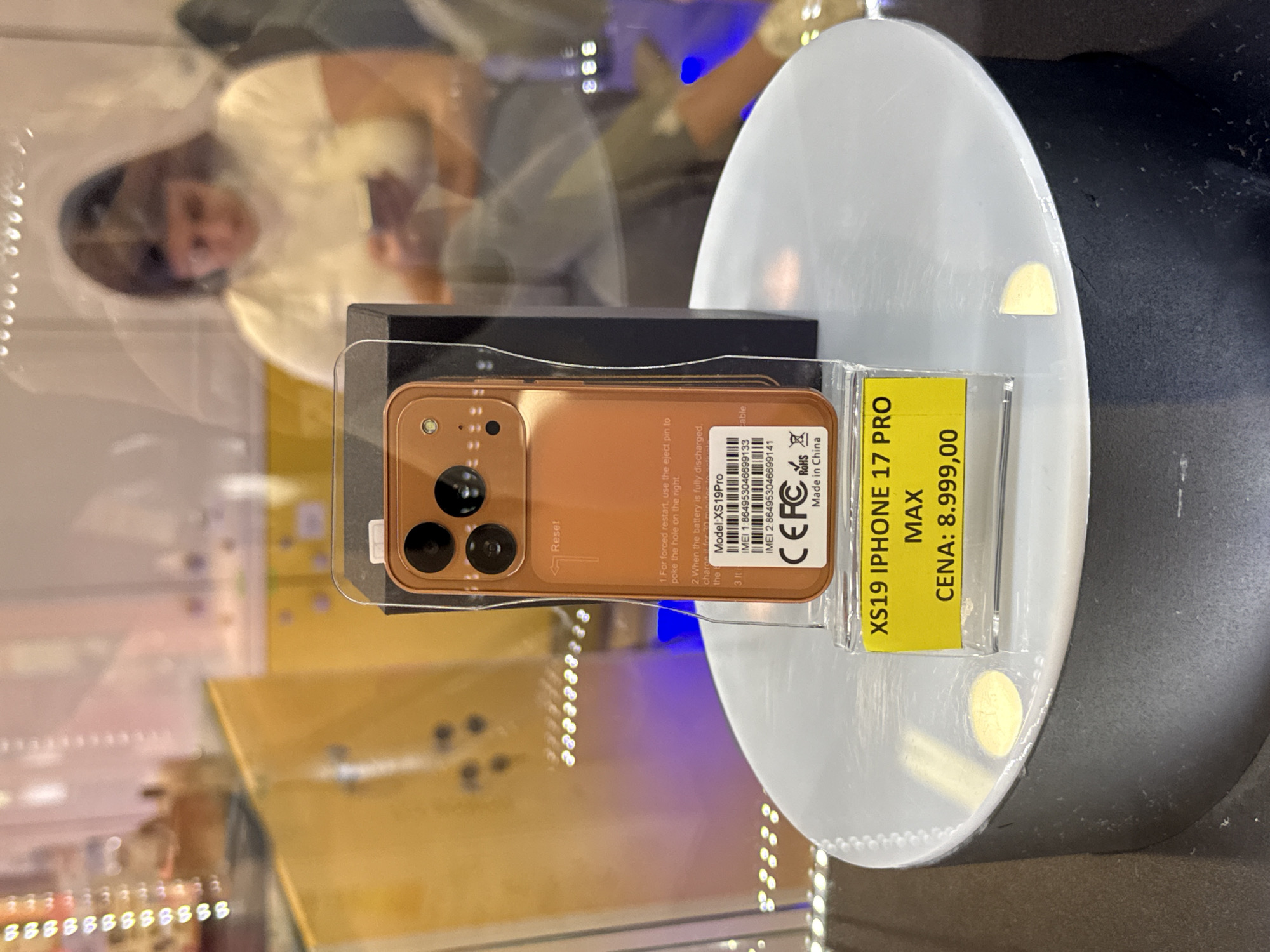 This image captures a retail display, likely inside an electronics store in Kragujevac, Serbia. The primary subject is a copper-gold colored smartphone, presented on a clear plastic stand atop a round, white display base. The phone is in packaging and appears to be a replica or generic model, as indicated by the "XS19 IPHONE 17 PRO MAX" text on a yellow price tag attached to the display stand.
Notable details include the phone's design, featuring a prominent triple-camera system on its back, typical of modern high-end smartphones. Text visible on the phone's back includes usage instructions such as "1. For forced restart, use the eject pin to poke the hole on the right," and partial instructions for charging. The model is specified as "XS19Pro," along with two IMEI numbers, "CEFCR" and "RoHS" compliance marks, and "Made in China." The price tag clearly states "CENA: 8.999,00," which, given the location, is likely in Serbian Dinars. A blurred reflection of a person, possibly a woman with dark hair, is visible in the background, suggesting an indoor, brightly lit environment. The overall scene depicts a product showcase intended for sale.