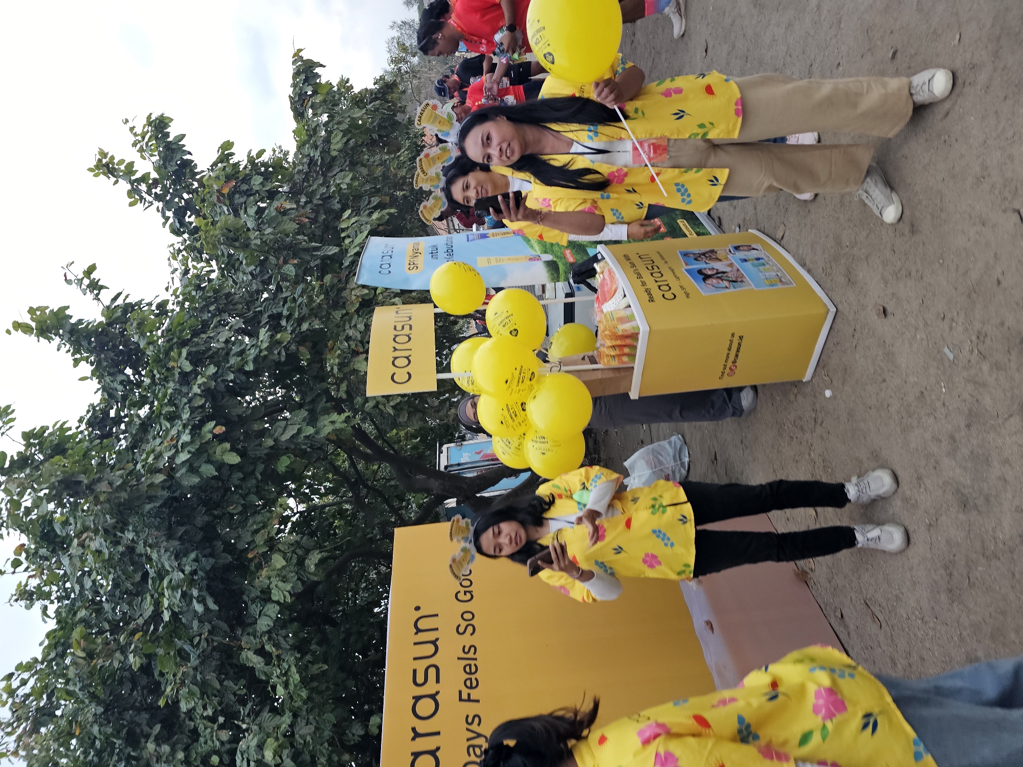 This outdoor scene in Denpasar, Indonesia, features a promotional event for "Carasun." In the foreground, two women are the primary subjects. The woman on the right, with long dark hair, wears a bright yellow floral-patterned shirt and tan pants, holding a white stick and a large yellow balloon. She is looking towards the camera. To her left, another woman, also wearing a similar yellow floral shirt and black pants, is looking down at her phone. In the background, several other people are visible, many wearing yellow, suggesting a coordinated event. Behind the two main subjects is a yellow promotional stand with "Carasun" branding and images of people. Several yellow balloons are clustered around the stand, adding to the festive atmosphere. A large green tree with dense foliage dominates the upper portion of the image, with a pale, overcast sky above. The ground appears to be dusty or unpaved, consistent with an outdoor event space in Denpasar.