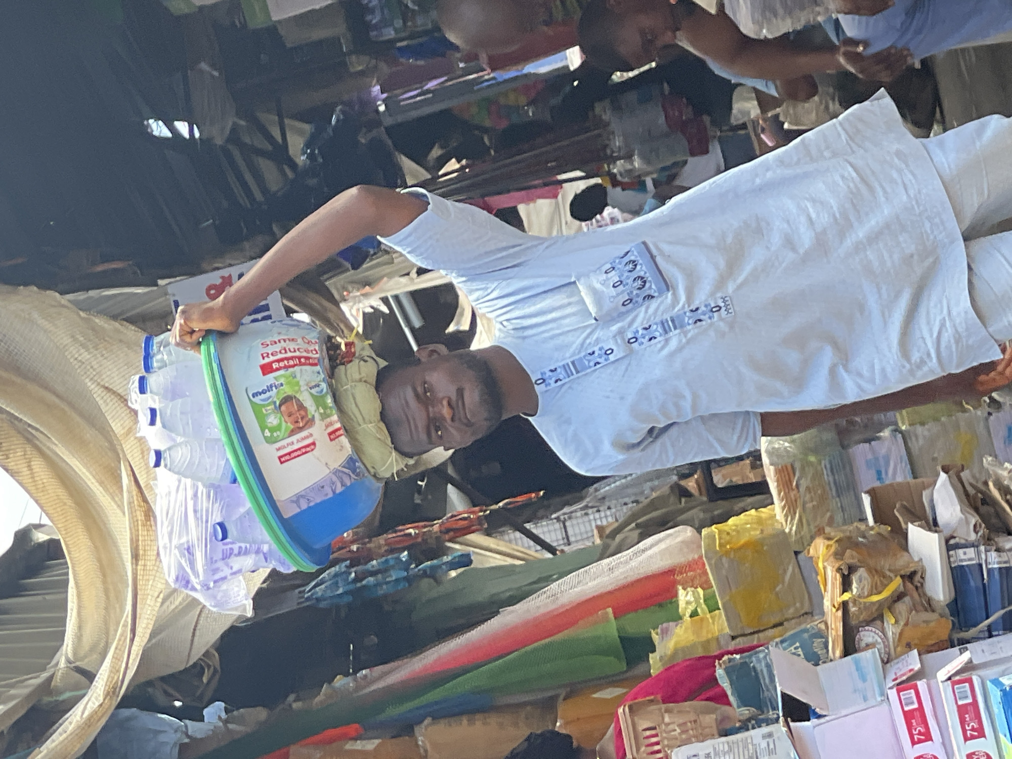 The image shows a man in a white traditional garment carrying a bucket of water bottles on his head in an outdoor marketplace. The bucket is blue and green, with a label for "Molfix" diapers. The surrounding area is cluttered with various goods for sale, including fabrics, packaged items, and boxes. The location is likely a market in Abuja, Nigeria, based on the visual cues of the environment and the style of clothing. Two other men are visible in the background.