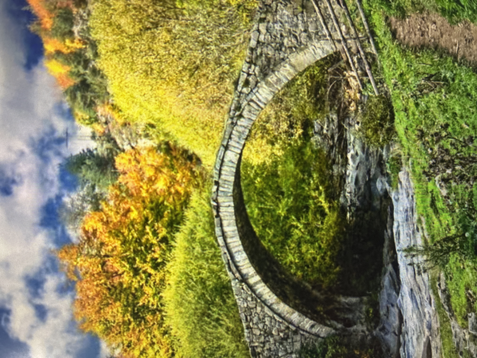 The image depicts a historic stone arch bridge spanning a deep, verdant ravine, characteristic of the mountainous terrain near Asenovgrad, Bulgaria. The bridge is constructed from rough-hewn grey stones, forming a classic, elegant arch. A simple, weathered wooden railing can be seen along one side of the bridge.
The surrounding landscape is densely forested, showcasing a vibrant blend of natural colors. The foliage includes bright greens, intermingled with striking yellows and oranges, suggesting an autumn season. Below the bridge, the ravine slopes are covered in dark green vegetation and rugged rock faces.
The sky in the background is a clear blue with scattered white clouds, indicating a bright, pleasant day. The scene conveys a sense of tranquility and natural beauty. There are no people visible, and no specific activity is taking place other than the static display of the landscape. No visible text is present in the image.