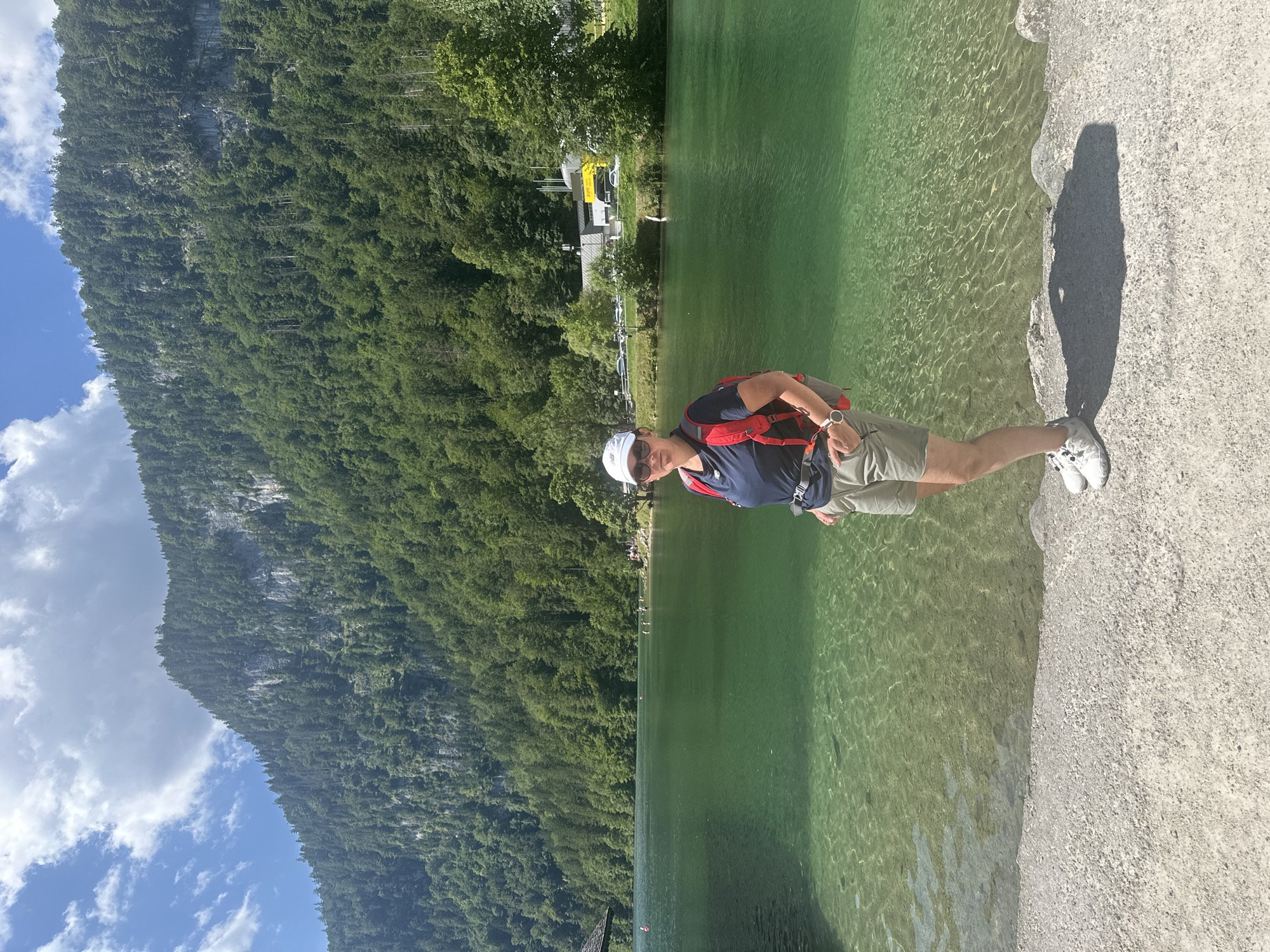 In the city of Schönau am Königssee, Germany, a woman stands on the shore of a crystal-clear green lake, with a lush green mountain forest serving as the backdrop. The woman, wearing a white cap, sunglasses, a dark blue t-shirt, khaki shorts, and white sneakers, has a red backpack on. The water is so clear that the lakebed is visible, with ripples creating a shimmering effect on the surface. The mountain slope is densely covered with trees, and a few buildings can be seen in the distance on the right side of the image, near the shoreline. The sky above is partly cloudy with a bright blue expanse.