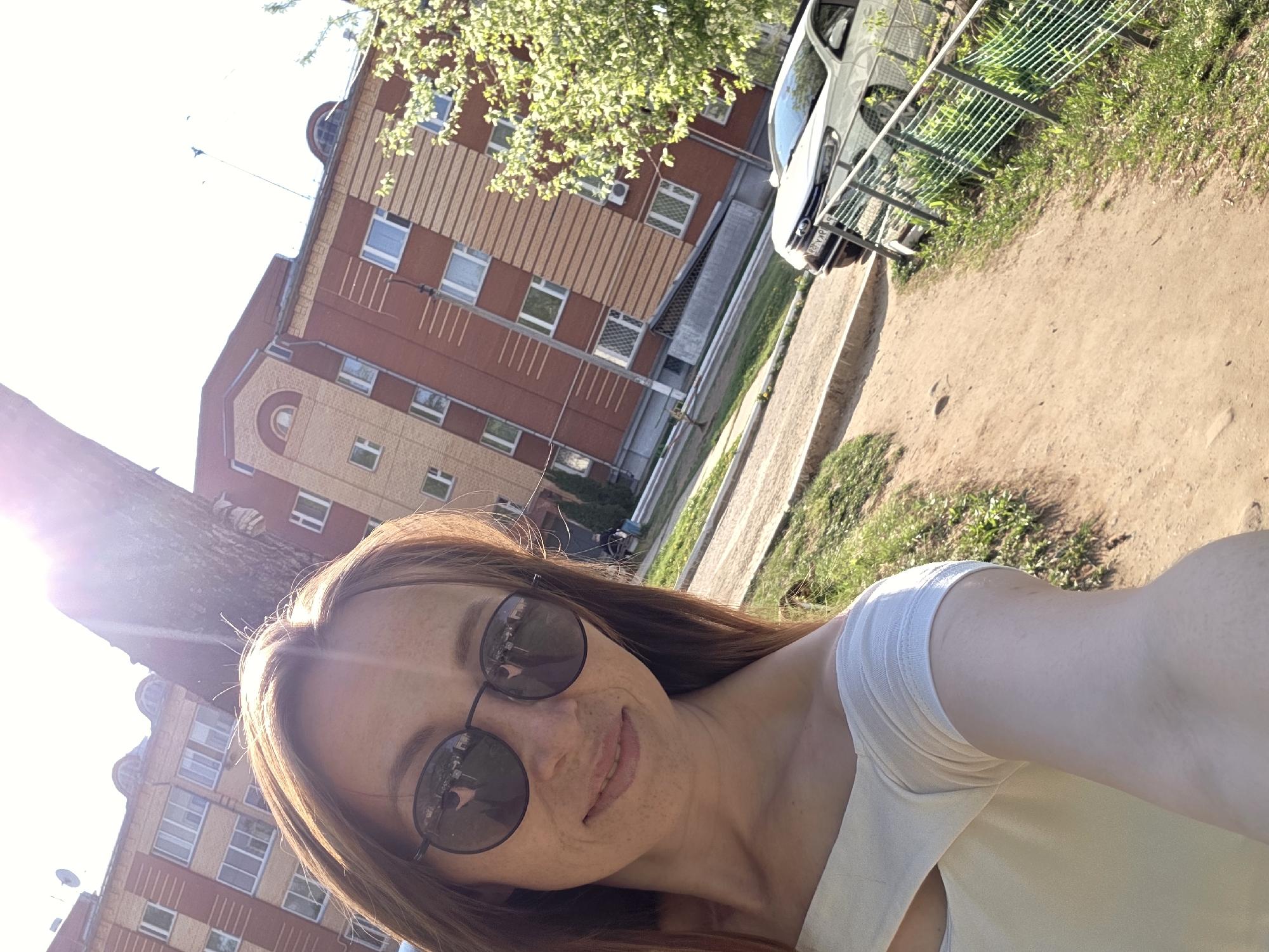 A selfie captured outdoors in daylight depicts a woman with long brown hair and round sunglasses. She wears a white top. Behind her is a tan-colored dirt path and a residential building with brick and tan paneling. The building is likely located in a Russian city, based on architectural styles and vehicles parked nearby. A white van is visible beside a green mesh fence. Branches of a tree are in the top left of the frame, near the woman's face. There is sunlight streaming into the left side of the photograph.