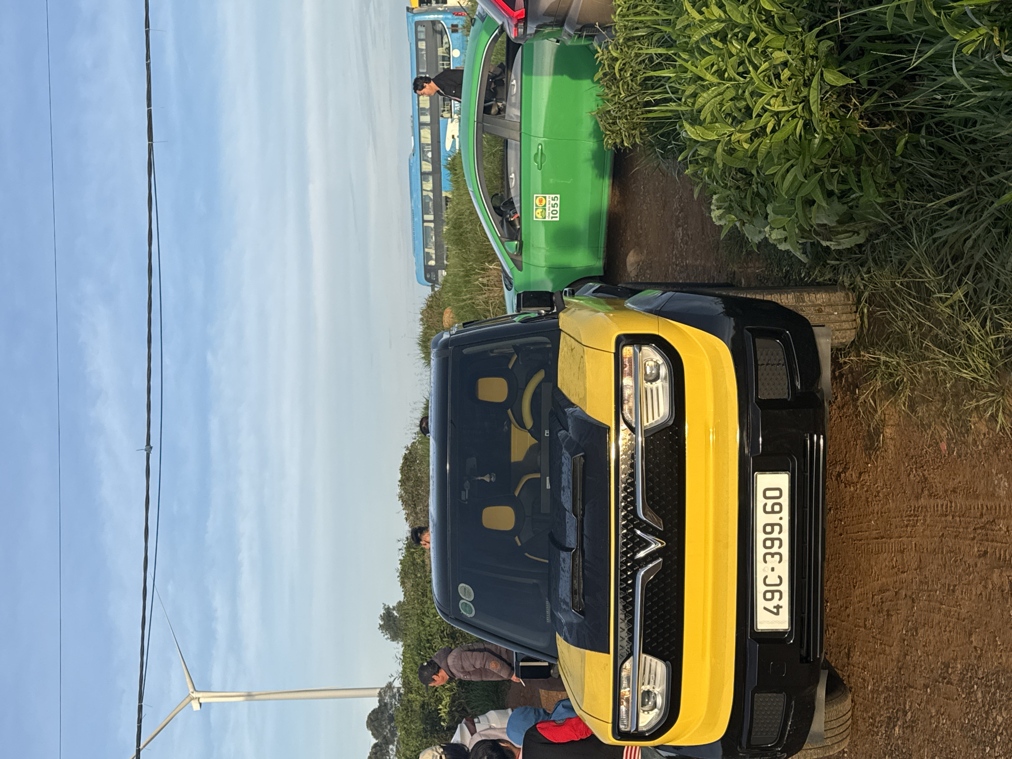 In Phát Chi, Vietnam, a yellow and black compact SUV is parked on a dirt road, facing the viewer. The SUV has a distinctive design with a V-shaped grille and horizontal headlights. A license plate on the front bumper reads "49C-399.60". To the right of the SUV, a green sedan is parked, with a taxi identification sticker and the number "1056" on its door. Further in the background, a long blue bus is visible, as well as other vehicles and people. The environment appears to be a rural or semi-rural setting, with grass and foliage to the right of the vehicles. The sky is blue with scattered clouds, and a utility pole with wires is present on the left side of the image.
