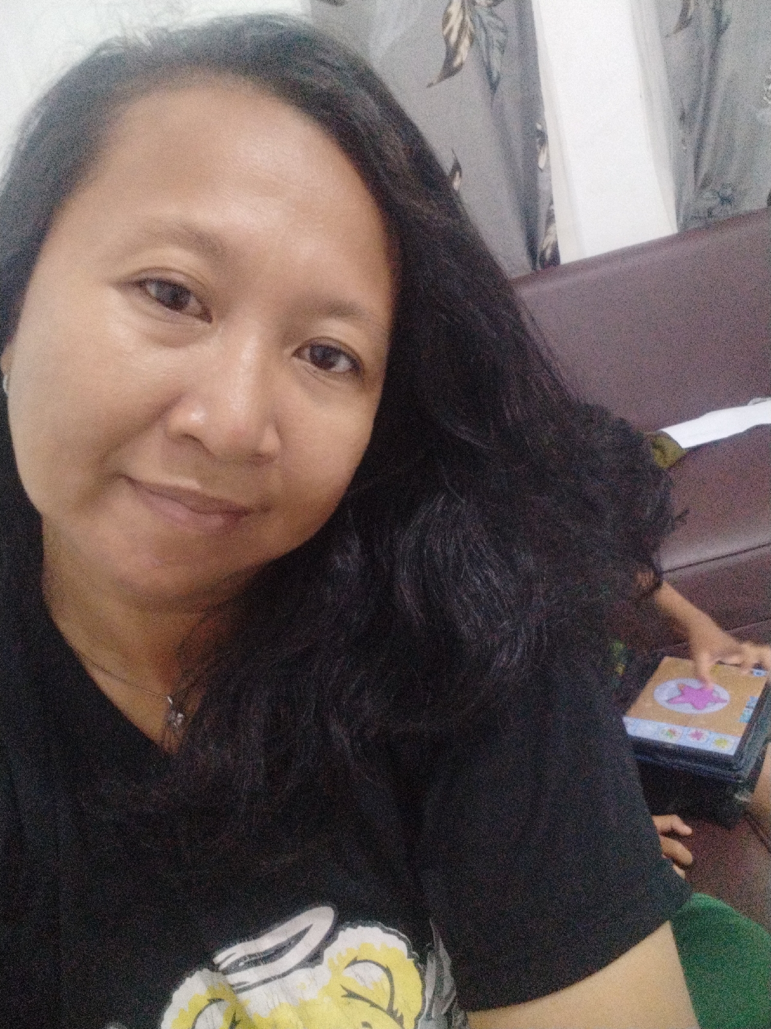 An adult with dark hair and medium skin tone is looking at the camera, possibly taking a selfie. They are wearing a black t-shirt with a yellow and white graphic. Behind them, a brown couch is visible, along with curtains with a floral pattern. A child's arm is also in the frame, reaching towards a stack of books. The scene appears to be indoors.