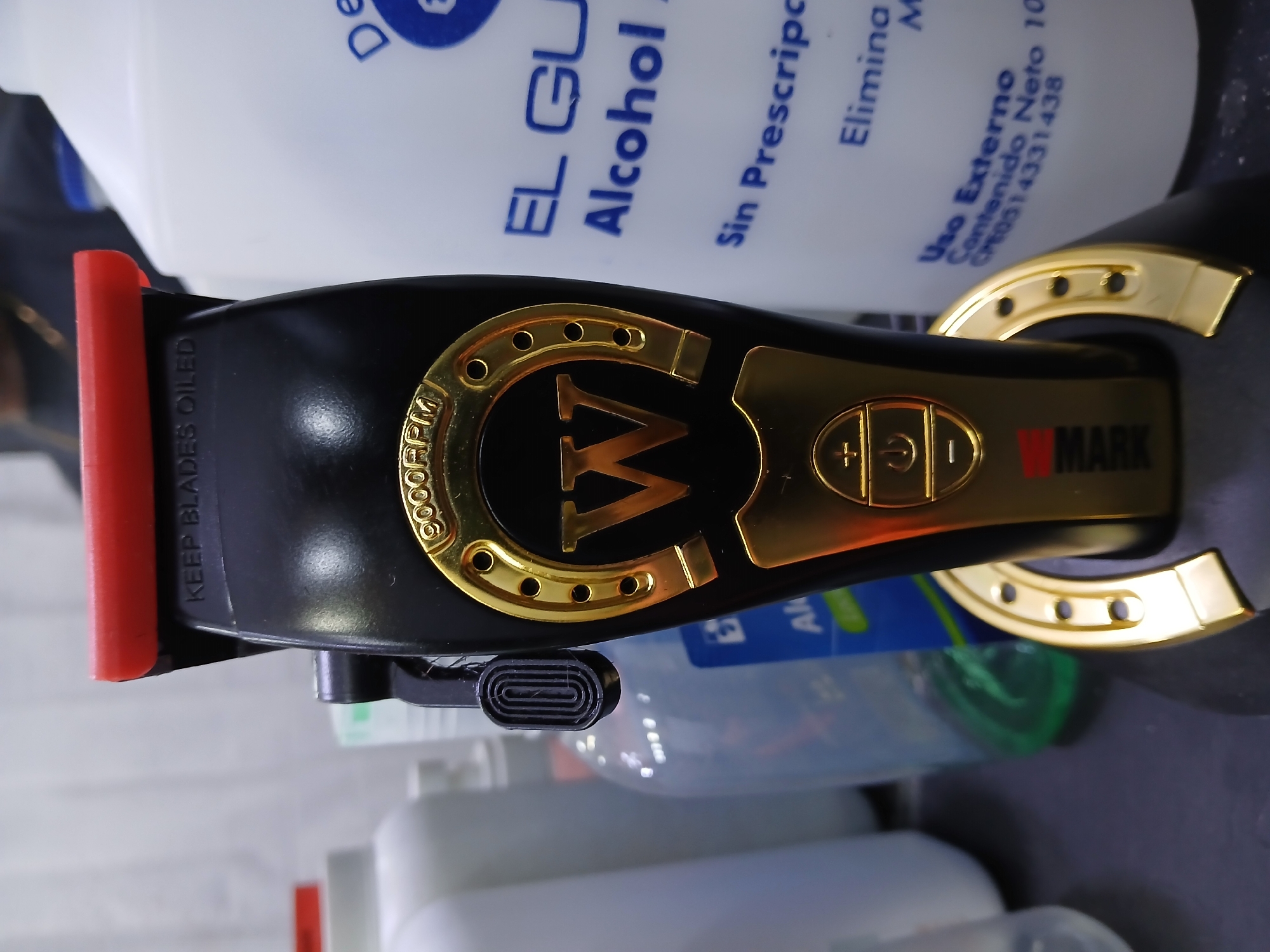 A close-up shot depicts a black and gold V-MARK hair clipper positioned near bottles of "EL GU Alcohol". The clipper has a red attachment at the top. Text on the clipper reads "KEEP BLADES OILED" and "9000RPM." The alcohol bottles are labeled in Spanish, "Sin Prescripc" and "Uso Externo." The overall setting appears to be a tabletop or grooming area. The location of Juan Griego, Venezuela is not evident in the image.