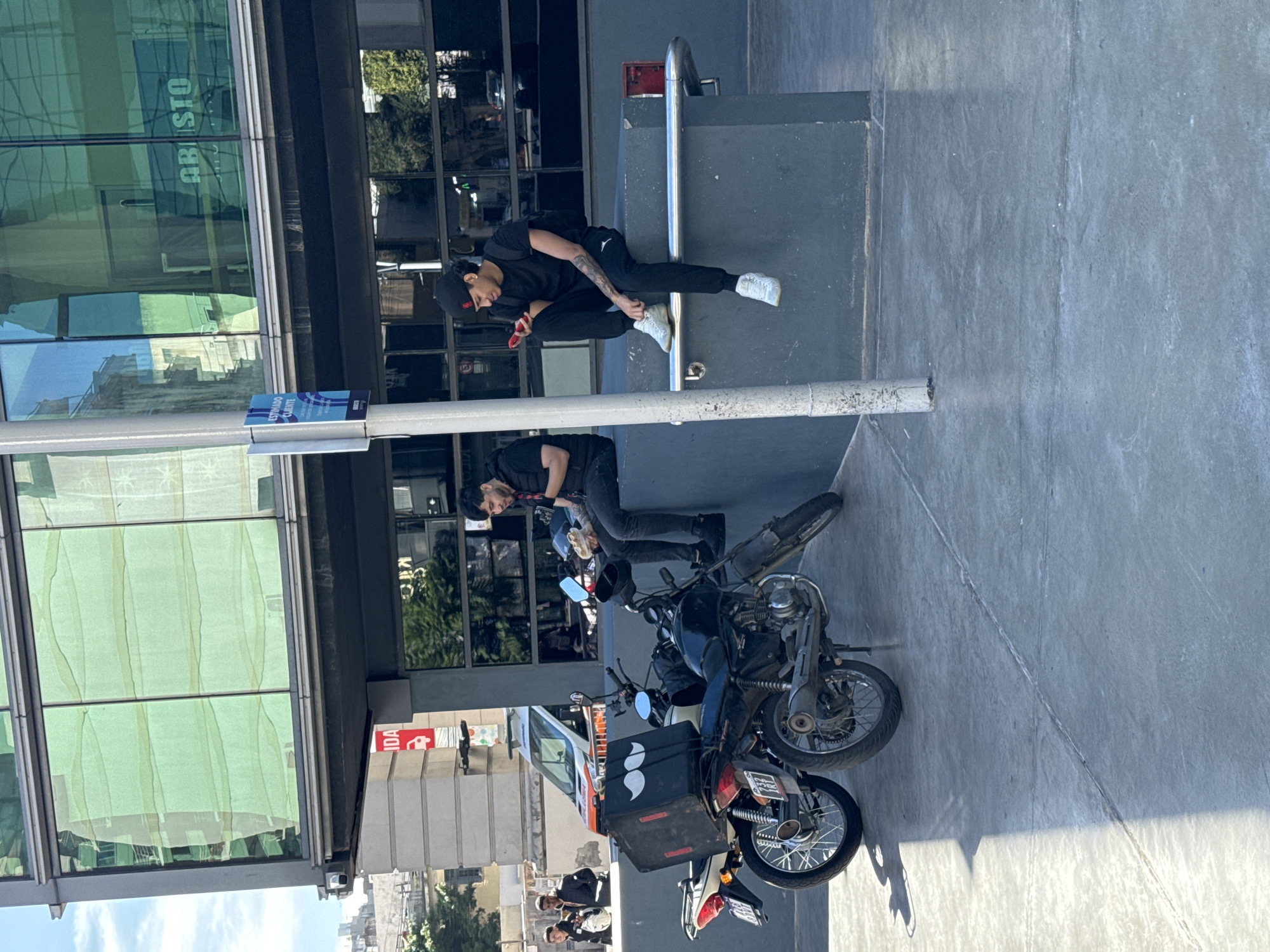 The image depicts an urban scene in Buenos Aires, Argentina. Two men are present; one is seated on a step in front of a building, using a mobile device, wearing a black cap, t-shirt, pants and white sneakers. The second man stands beside two black motorcycles, and wears similar apparel: black cap, t-shirt, and pants. A delivery box is attached to one of the motorcycles, adorned with a white mustache logo. The environment consists of a modern building with a reflective glass facade. The architecture includes a sign displaying the word "ABASTO." The setting includes urban elements, such as a sidewalk, a metal handrail, and street-level infrastructure.