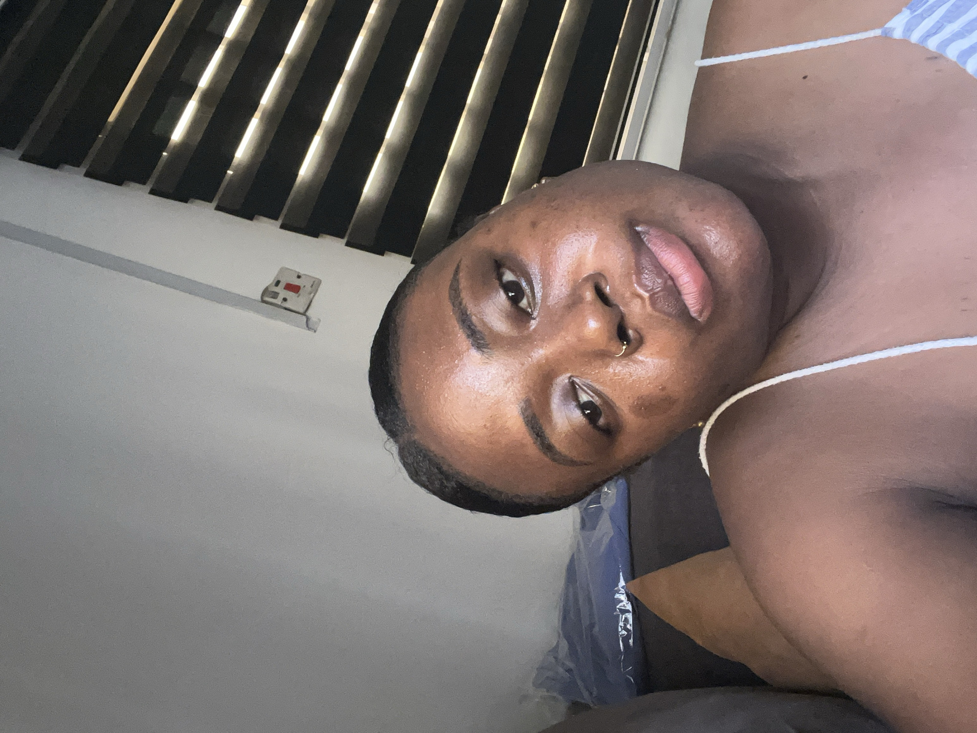 A medium shot of a dark-skinned person with a gold nose ring is angled, with a bedroom window showing drawn blinds in the upper-left corner. The person is wearing a white string-like strap, and a blue striped pattern is partially visible near the right shoulder. A light switch is visible on the white wall near the blinds. The background includes blue plastic, an arm and clothing. The location is likely indoors, possibly in Abuja, Nigeria.