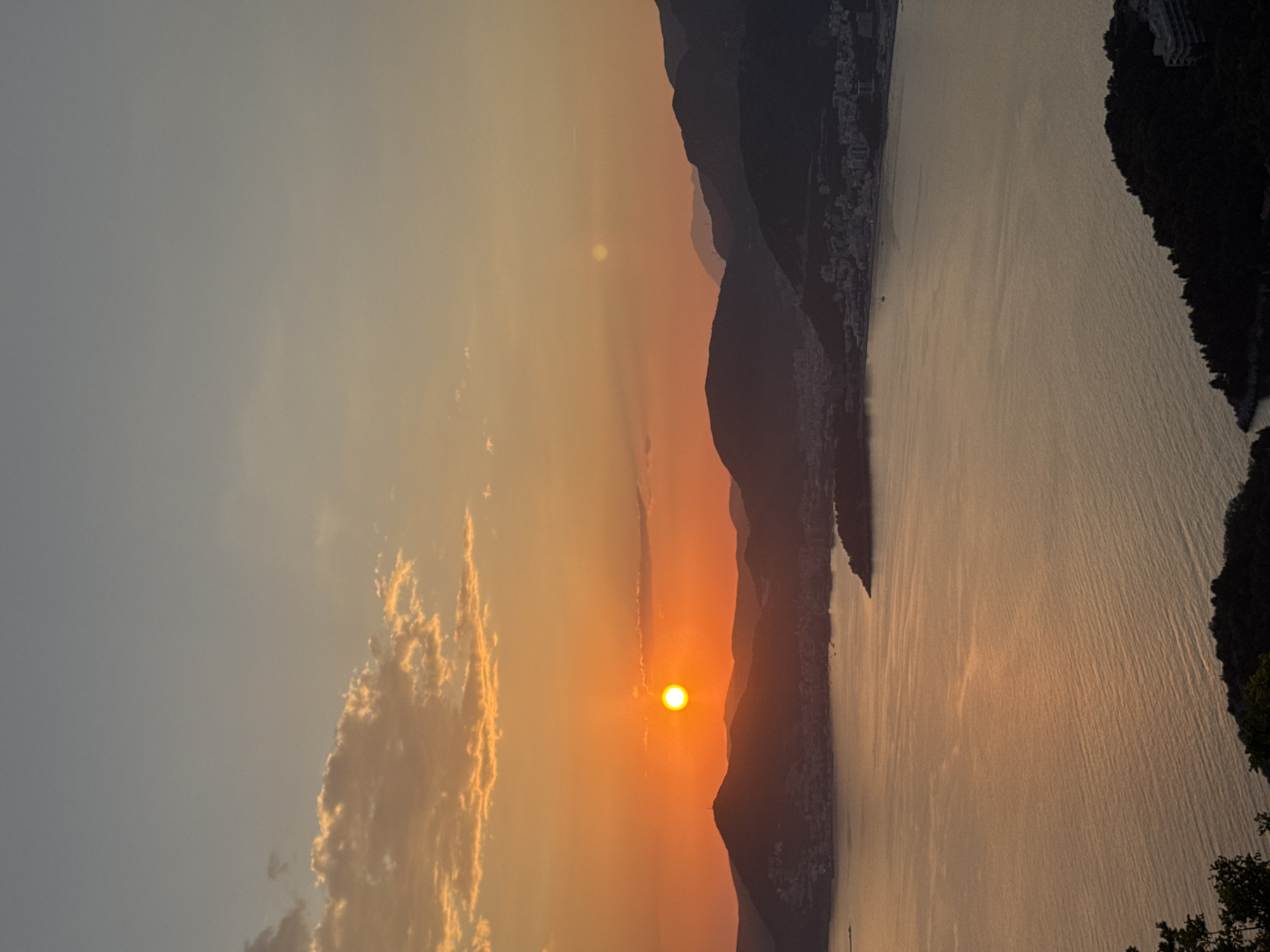 The image depicts a sunset over the Adriatic Sea, likely taken near Sveti Stefan, Montenegro, given the presence of mountainous terrain and the coastal setting. The sun, appearing as a bright orange orb, is situated near the horizon, partially obscured by a series of dark mountain silhouettes. The sky transitions from a pale blue at the top to a vibrant orange near the horizon. Fluffy, golden-tinged clouds are visible in the lower left of the frame. The water reflects the colors of the sunset, creating a shimmering effect on the surface. Vegetation is visible on the right edge of the frame. A few buildings are visible on the left shore.