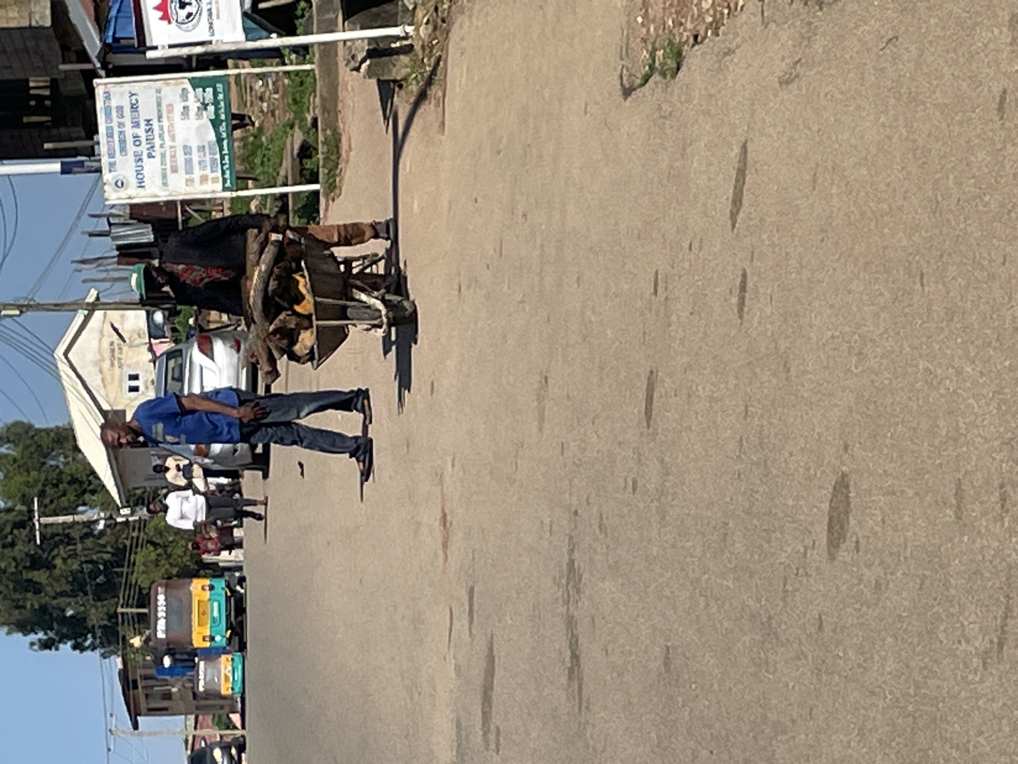The image depicts a street scene in Jos, Nigeria, under a clear sky. In the foreground, a man wearing a dark jacket, a patterned scarf around his neck, and a green cap is pushing a wheelbarrow filled with firewood. He is walking on a paved road. To his left and slightly behind him, another man in a blue t-shirt and jeans is walking.
In the background, several vehicles are visible on the road. These include a small yellow and green passenger vehicle resembling a tuk-tuk, and a larger bus with the registration number "PRD 5550". Further down the road, a silver car is parked.
A white sign with text is mounted on a pole to the right of the man with the wheelbarrow. The sign has a logo at the top and appears to be an informational or directional sign. Buildings line the street, with some featuring corrugated metal roofs. Trees are also present, indicating a partially urbanized or suburban environment. The ground is mostly paved road with some patches of dirt and vegetation on the sides. The overall impression is that of a typical day in a Nigerian city, with people engaged in daily activities.