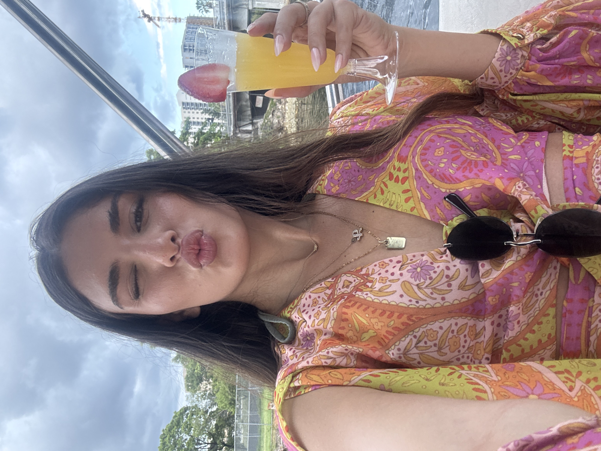 A woman with long, dark hair and fair skin poses for a selfie on what appears to be a boat, holding a drink with a strawberry garnish. She is winking and puckering her lips, wearing a colorful, patterned blouse. The background shows a cloudy sky, trees, buildings, and a bridge, suggesting an urban waterfront location in Miami, Florida.