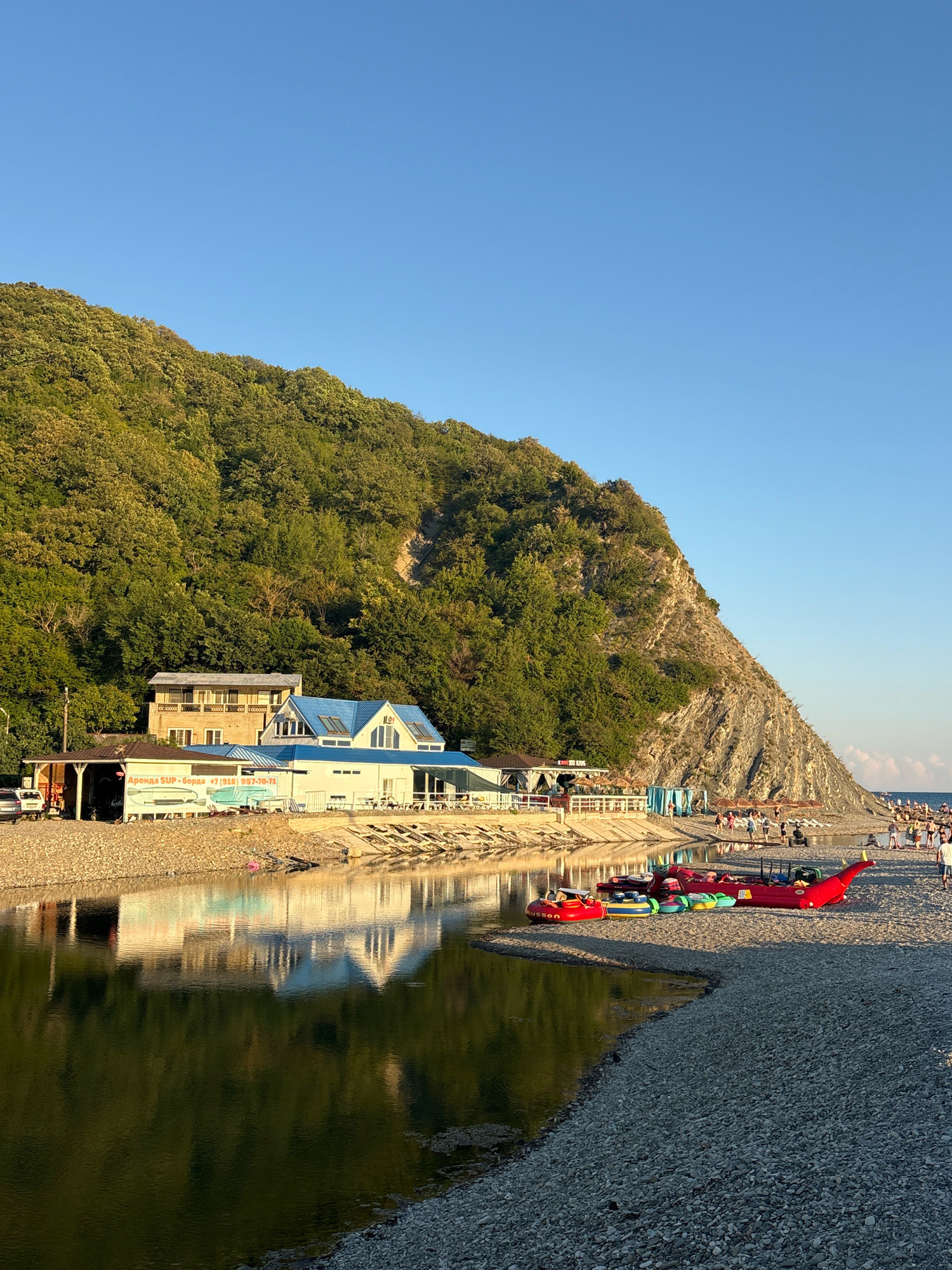 The image depicts a scenic view of the coastline in Arkhipo-Osipovka, Russia, characterized by a steep, green, tree-covered mountain slope on the left, dropping down to a rocky beach. A tranquil body of water, likely a river or inlet, separates the beach from a strip of buildings situated at the base of the mountain.
The buildings appear to be part of a resort or recreational area, with one prominent structure featuring a bright blue roof. Signs on the buildings indicate commercial activity, with text in Cyrillic that includes "Аренда SUP - борда" (Rent SUP - board) and a phone number. Several small boats, including inflatable kayaks and a larger red inflatable vessel resembling a dragon, are arranged on the pebble beach near the water's edge. Further along the beach, to the right, numerous people are visible, some lounging under umbrellas and others walking. The water reflects the blue sky and the buildings. The sky is clear and a vibrant blue, indicating fair weather, and the low angle of the sun suggests it is either early morning or late afternoon, casting a warm light on the scene. The terrain is predominantly composed of smooth, gray pebbles.