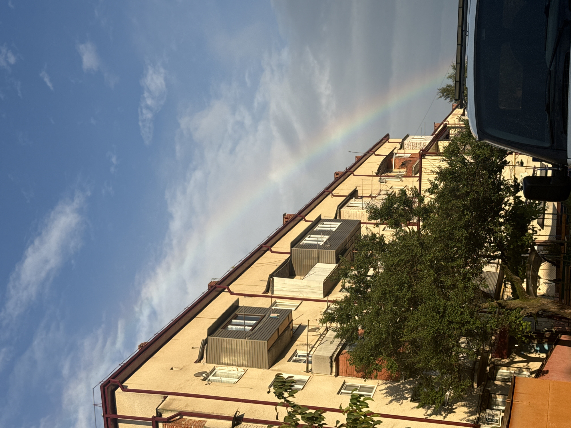 In Beslan, Russia, a multi-story apartment building with a beige facade and red trim stands under a partially cloudy blue sky. A faint rainbow arcs across the sky, visible over the building. Several balconies extend from the building, some enclosed with grey paneling and others open with white railings. A leafy green tree is in the foreground, casting shadows on the building. A portion of a white vehicle, likely a van, is visible in the lower right corner of the image, its windshield slightly wet. The scene suggests a post-rain moment with the sun breaking through.