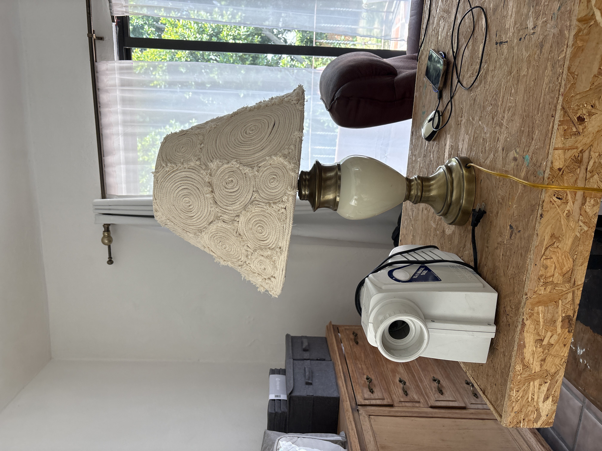 The image shows an indoor scene with a window, a lamp, a projector, and furniture.
A window with white sheer curtains is in the background, revealing trees outside.
A lamp with a tan-colored lampshade decorated with circular patterns sits on a wooden surface. The lamp has a gold and white base.
A white projector is positioned near the lamp on what appears to be a wooden cabinet or table.
The background includes a brown upholstered sofa or chair. There is a wooden structure behind the lamp with a computer mouse and a phone.
The scene is undated. The image does not provide sufficient information to identify the precise location in Puebla, Mexico.