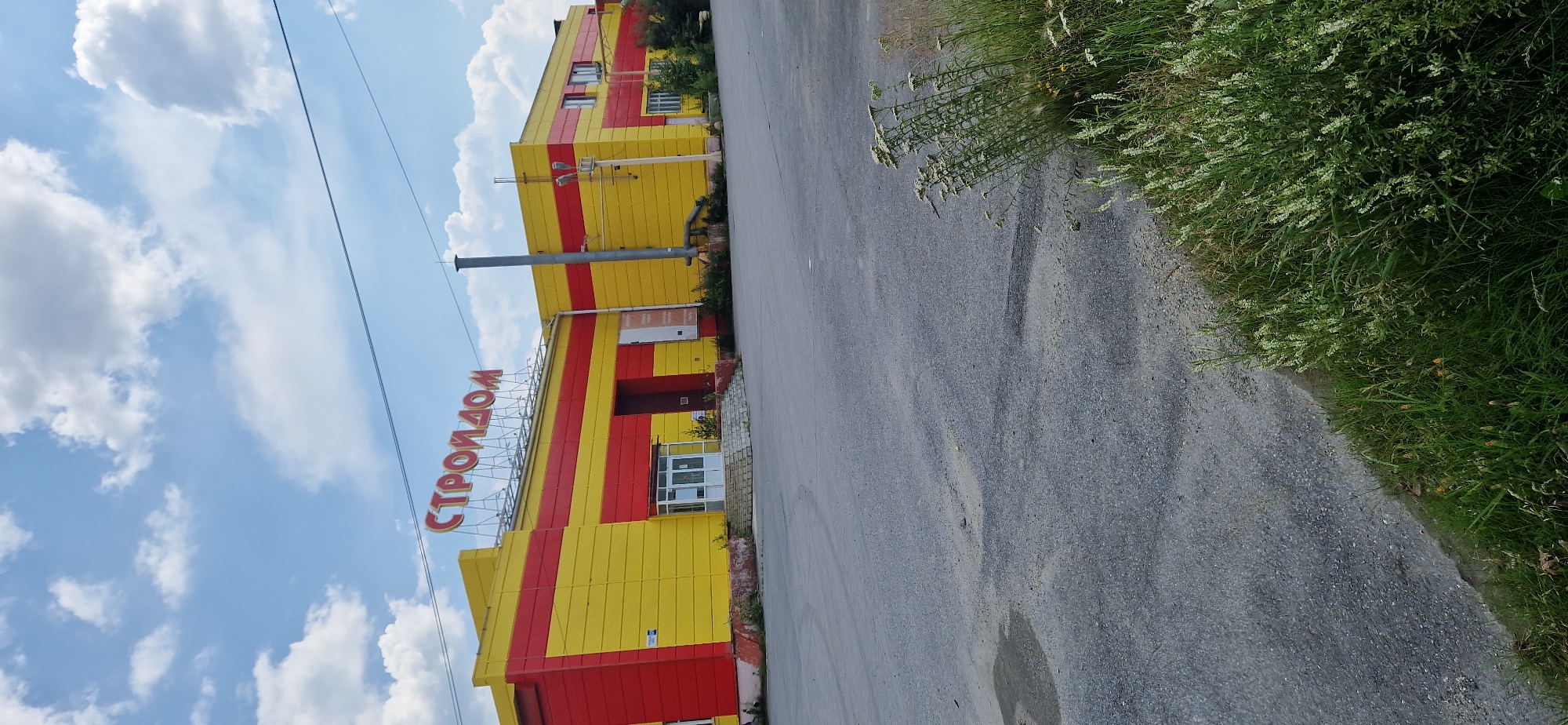 An exterior view shows a yellow and red building with the word "СТРОЙДОМ" in large red letters on its facade, under a blue sky with white clouds. The building is a commercial structure with large, roll-up doors and several windows. A paved lot with some patchy vegetation and dry grass is in the foreground. The building appears to be a retail store or warehouse, possibly for building materials given the signage. The scene suggests a location in a city, with infrastructure like power lines visible. Based on the Cyrillic text and architectural style, this building is likely located in Miass, Russia.