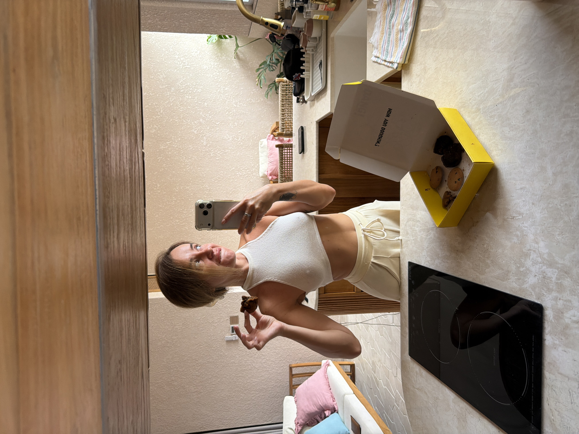 A woman in a white crop top and cream-colored pants is taking a selfie in a kitchen reflected in a mirror. She holds a cookie in her right hand and her phone in her left. A yellow box of cookies sits open on the counter in front of her. The kitchen appears modern with wooden cabinets, a sink with a gold faucet, and a stovetop. There is also a couch with pink and blue pillows visible in the reflection. The woman's hair is pulled back, and she is looking to the side with a slight smile. The location is identified as Mengwi, Indonesia.