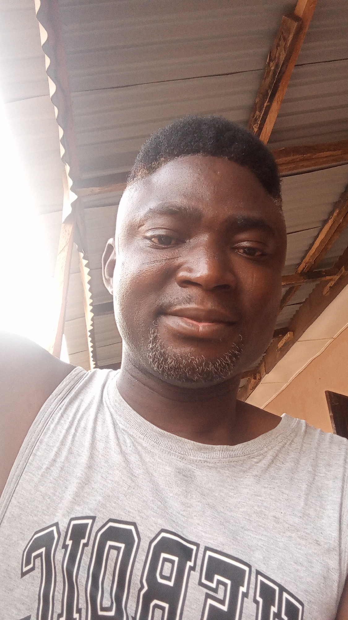 A close-up selfie of a dark-skinned man wearing a gray tank top with black lettering. The man has short black hair and a graying goatee. The background shows a corrugated metal roof with wooden supports and part of a building with a doorway. The location is Sabon Tasha, Nigeria.