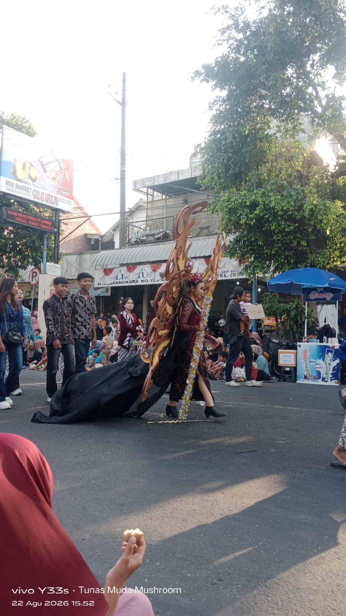 An outdoor street parade is underway in Genengan, Indonesia, with participants in traditional attire and elaborate costumes. In the foreground, a person wearing a burgundy hijab is partially visible, holding a small piece of food. The central focus is a performer in a detailed, wing-like costume made of gold and brown material, embellished with sparkling silver trim. This performer is walking on the asphalt street, trailing a large black fabric. To their left, three young men in patterned shirts stand by, observing the parade. Further back, several other individuals, including women in traditional Indonesian clothing and children, are gathered to watch. The street is lined with buildings, one of which has a sign indicating a business with "JL. LLAYANGKARA NO. 125" and a phone number. A blue umbrella, possibly advertising a product, is visible on the right side of the street. The environment suggests a sunny day, with shadows cast across the road and foliage from trees overhead. The timestamp "22 Agu 2026 15.52" and the text "vivo Y33s • Tunas Muda Mushroom" are present in the lower left corner, indicating the device and possibly the event or location.