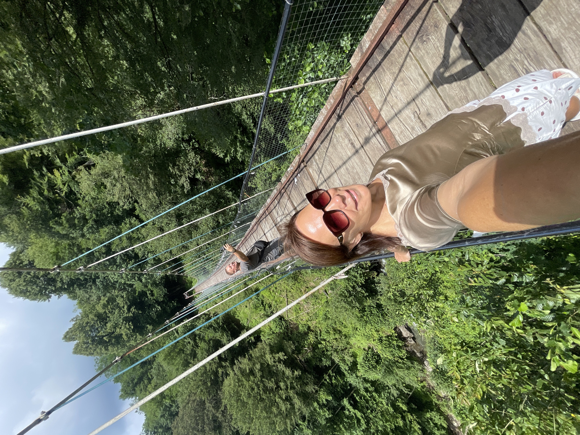 A woman wearing sunglasses and a silky top with white lace trim is taking a selfie on a wooden suspension bridge surrounded by lush green trees. In the background, a man in a grey t-shirt and shorts is also taking a photo. The bridge has metal railings and is suspended over a wooded ravine, with a glimpse of water below. The sun casts shadows on the wooden planks of the bridge. The scene appears to be in a natural, forested area.
