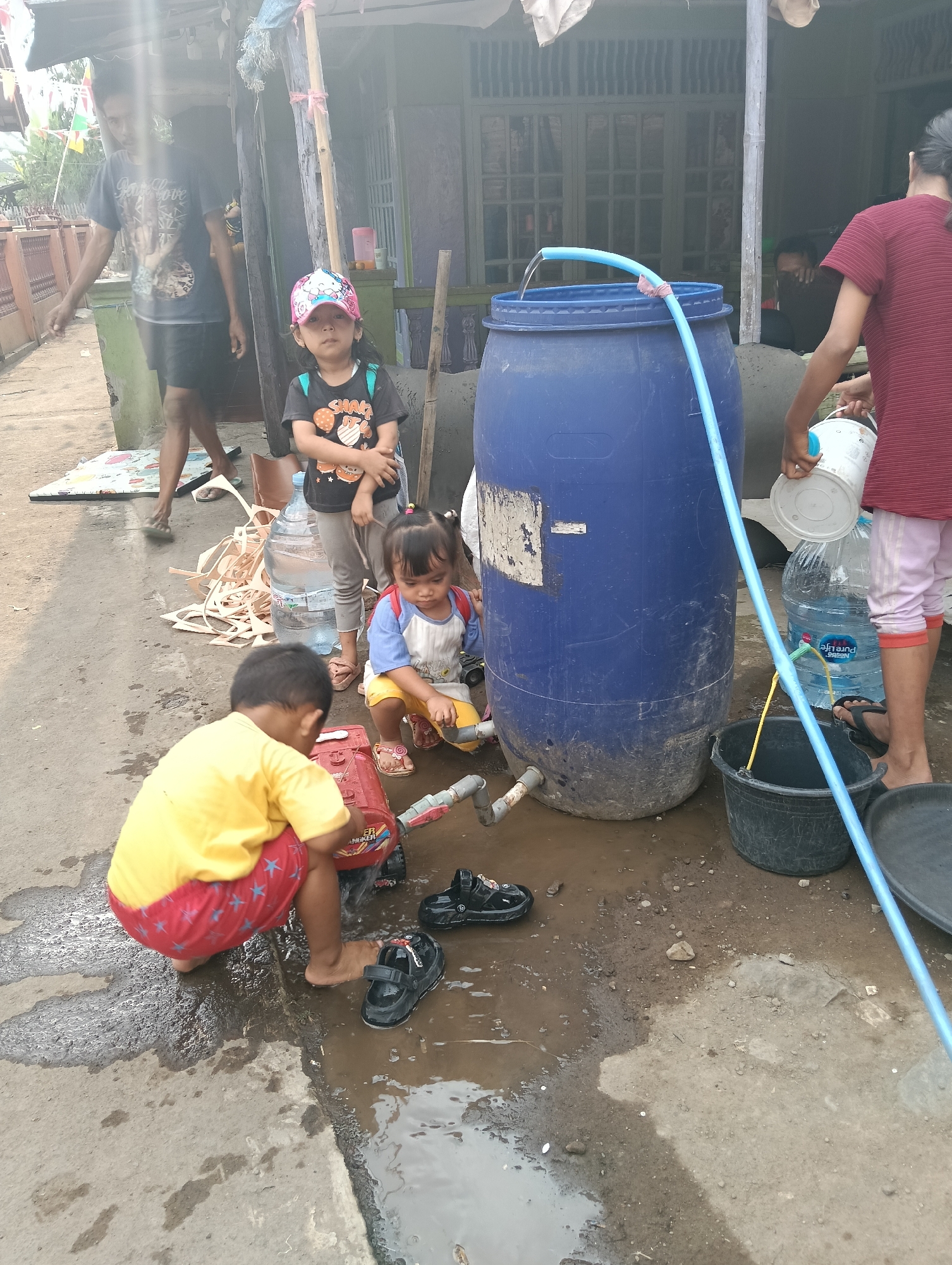 The image depicts an outdoor scene, likely in a residential area of Tasikmalaya, Indonesia, where children are interacting with water. In the foreground, a young boy wearing a yellow t-shirt and red shorts with white stars is kneeling on the wet ground, playing with a red toy car. To his right, a pair of black sandals lies in a puddle. Further back, a young girl wearing a blue and white shirt with yellow shorts is also seated and appears to be playing with water. She is wearing pink sandals. To her left, another child in a black t-shirt and grey pants and a pink and white cap stands with hands clasped. Behind them, a large blue plastic barrel is visible, with a blue hose running from it, seemingly dispensing water. A large bottle of "Nestlé Pure Life" drinking water is also present. To the right of the barrel, a person in a red striped shirt and pink pants is holding a white bucket, and a black bucket is situated on the ground. In the background, a man in a dark t-shirt and shorts stands, appearing to observe the scene. The environment suggests a humble dwelling with a visible structure and some debris on the ground, indicating a utilitarian outdoor space. The presence of puddles and the activity with water suggest recent or ongoing water collection or play.