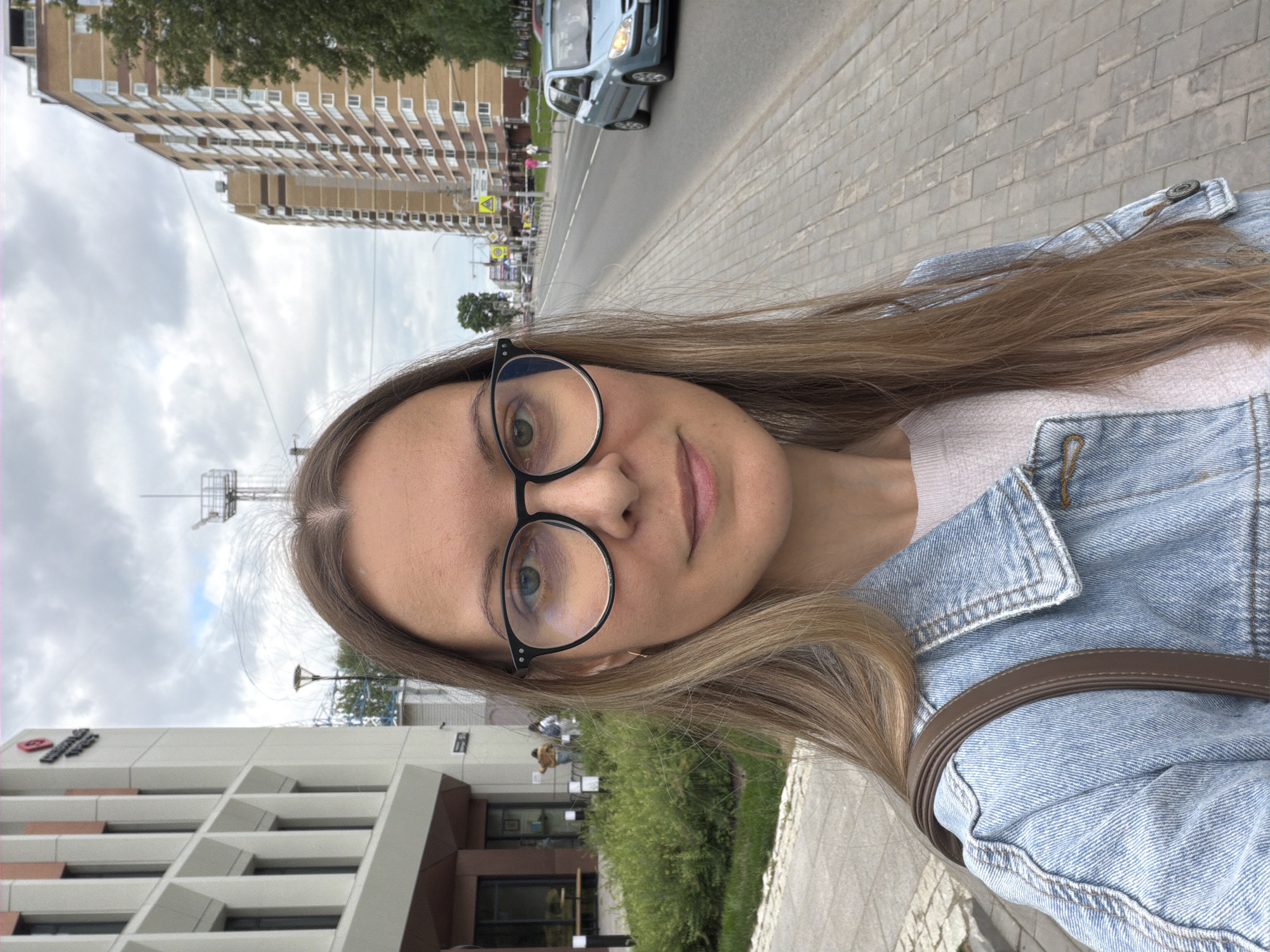 A woman is taking a selfie on a street in Izhevsk, Russia. She has long blonde hair, is wearing glasses, a light pink sweater, and a blue denim jacket. A brown purse strap is visible over her left shoulder. She is standing on a paved sidewalk next to a green grassy area. To her left is a modern, light-colored building with dark windows. To her right, across a road, are several multi-story apartment buildings and trees. A light blue car is driving on the road. The sky is cloudy.