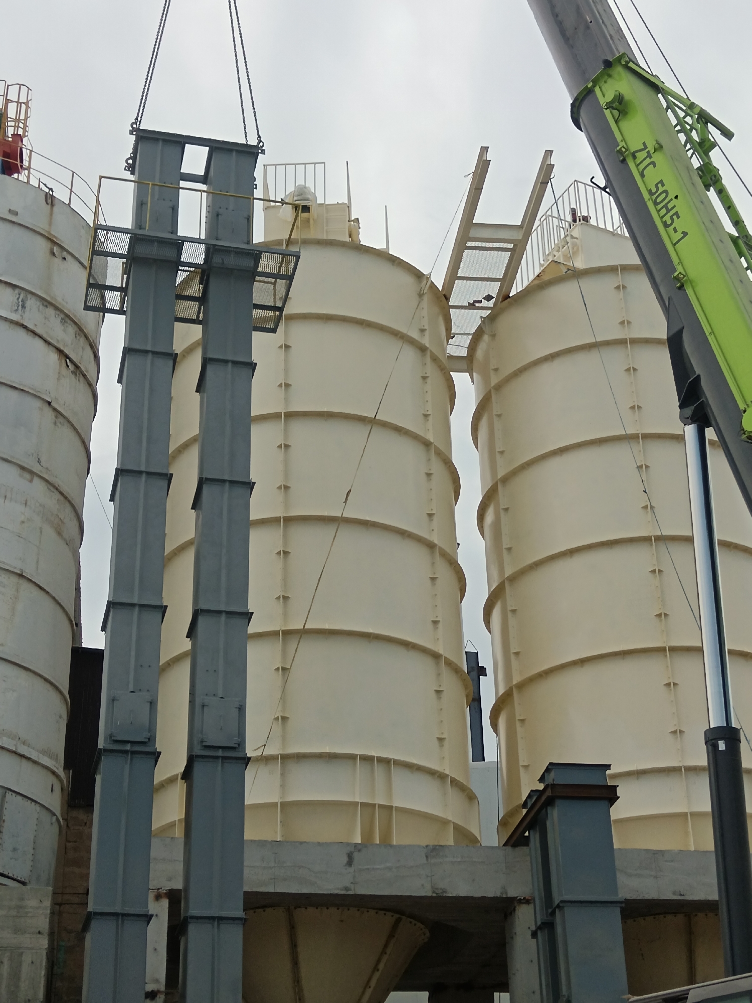 The image depicts an industrial scene in Aragatsavan, Armenia, focusing on the construction or maintenance of large, beige cylindrical silos. A large green crane, with the inscription "ZTC 50H5-1" visible on its boom, is actively engaged in lifting a grey, multi-sectioned metallic structure. This structure appears to be a vertical conduit or support system, suspended by heavy chains attached to the crane. Two identical beige silos dominate the background, with a third, partially visible white silo to the left. The silos are ribbed and appear to be made of metal. In the foreground, the grey metallic structure being lifted by the crane is positioned between the silos. The overall environment is overcast, suggesting cloudy weather. The scene is devoid of people, highlighting the mechanical nature of the operation.