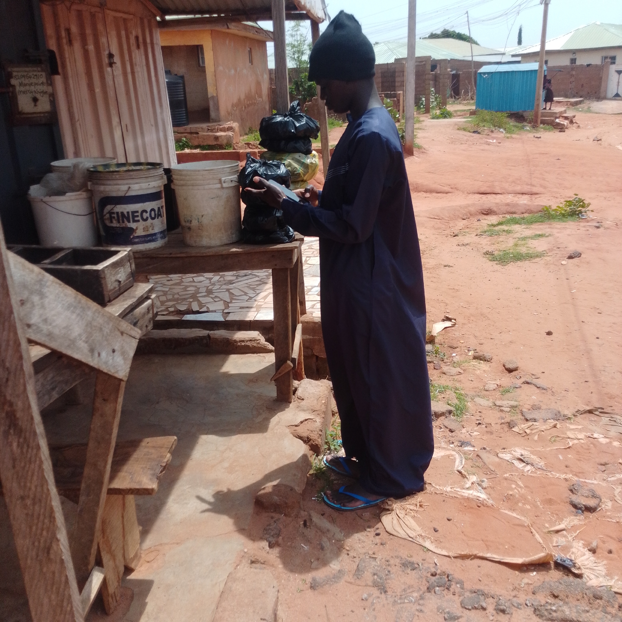 In Tumfure, Nigeria, a young person wearing a dark blue robe and a black beanie stands by a wooden table laden with various items. On the table, there are several buckets, including one labeled "FINECOAT." Piled on the table are multiple black plastic bags, some appearing full. The person is holding a mobile phone and looking down at it, suggesting they are engaged with the device. The environment is dusty and reddish, with visible debris and patches of grass on the ground. In the background, there are buildings with corrugated metal roofs and brick construction, typical of a semi-urban or rural setting in Nigeria. A bright blue corrugated metal structure is visible further in the background.