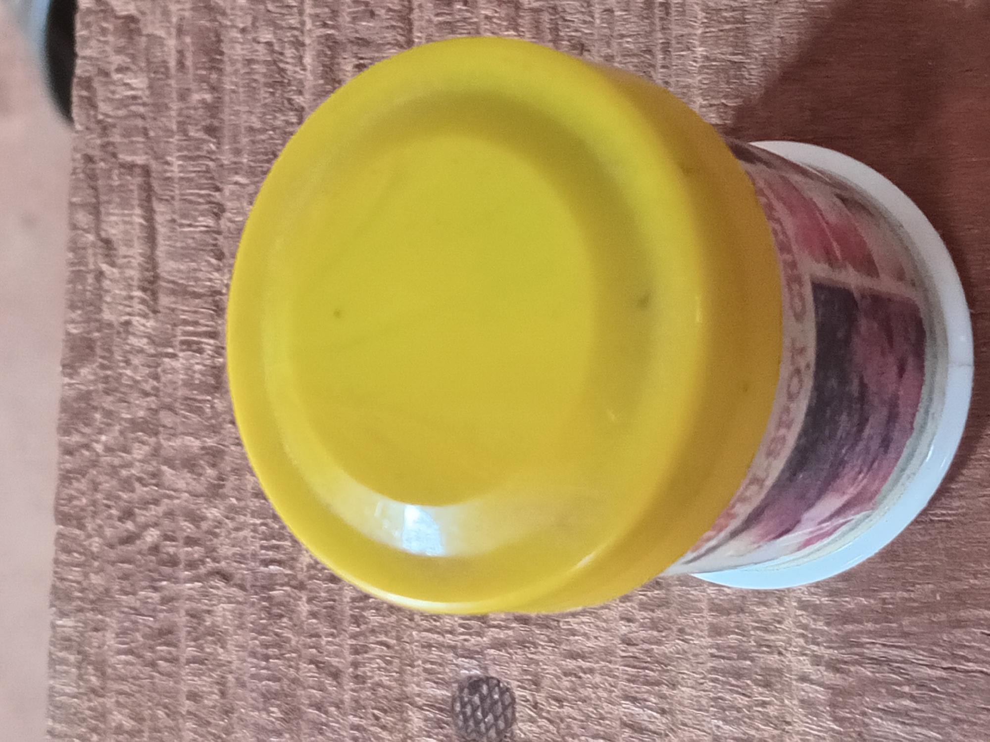 A close-up, rotated overhead shot displays a cylindrical container topped with a bright yellow, round plastic lid. The container rests on a textured, brownish-pink surface, likely wood or a similar material, characterized by parallel grooves and a somewhat rough appearance.
The yellow lid, smooth and slightly reflective, dominates the left side of the frame. Small dark blemishes or dust particles are visible on its surface. The container itself, visible below the lid on the right, is made of a light-colored material, possibly white plastic or paper. A colorful label wraps around its side, featuring abstract patterns and blocks of color including shades of red, pink, and dark purple or black. Partially visible text on the label appears to read "O-T SRO" or "T SPO" along with other indistinct characters.
On the textured surface, below the container, a small, dark, intricately textured object is visible, resembling a screw head or a decorative metal stud. The lighting is even, suggesting an indoor setting with diffused or overhead illumination, casting minimal shadows and creating a soft sheen on the yellow lid. No people are present, and no activity is occurring; the image is a static still life of the object.