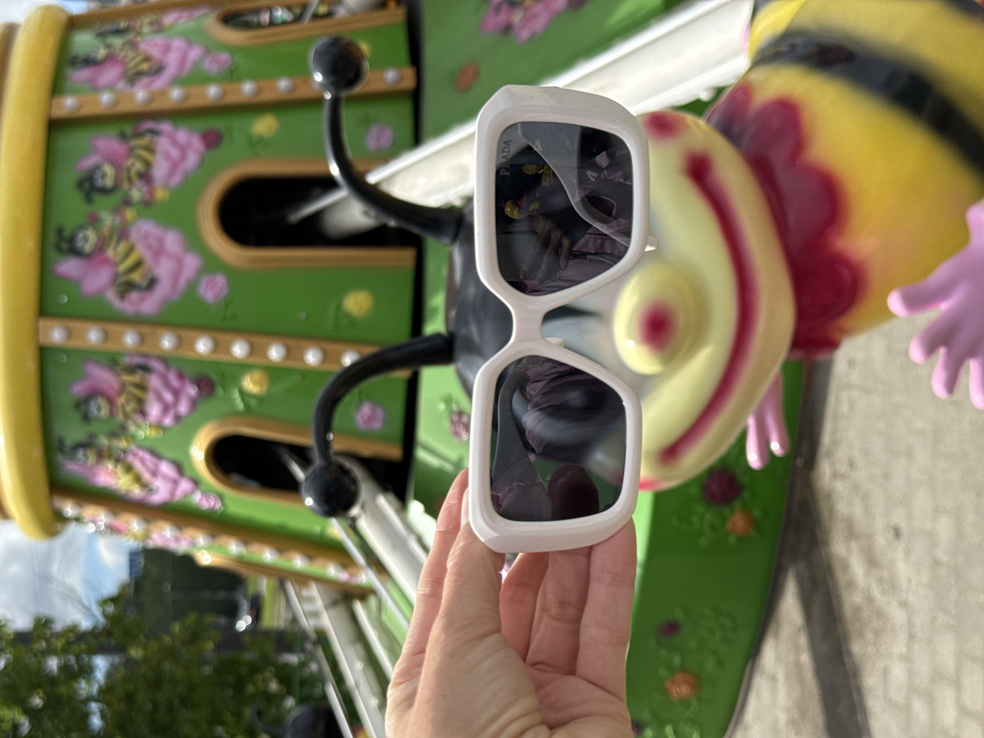 A hand holds a pair of Prada sunglasses in front of a brightly colored bee-themed amusement ride in Saint Petersburg, Russia. The ride is decorated with images of bees on flowers and has a yellow and green color scheme. A large bee figure is also present. The sunglasses reflect the ride and surrounding environment.