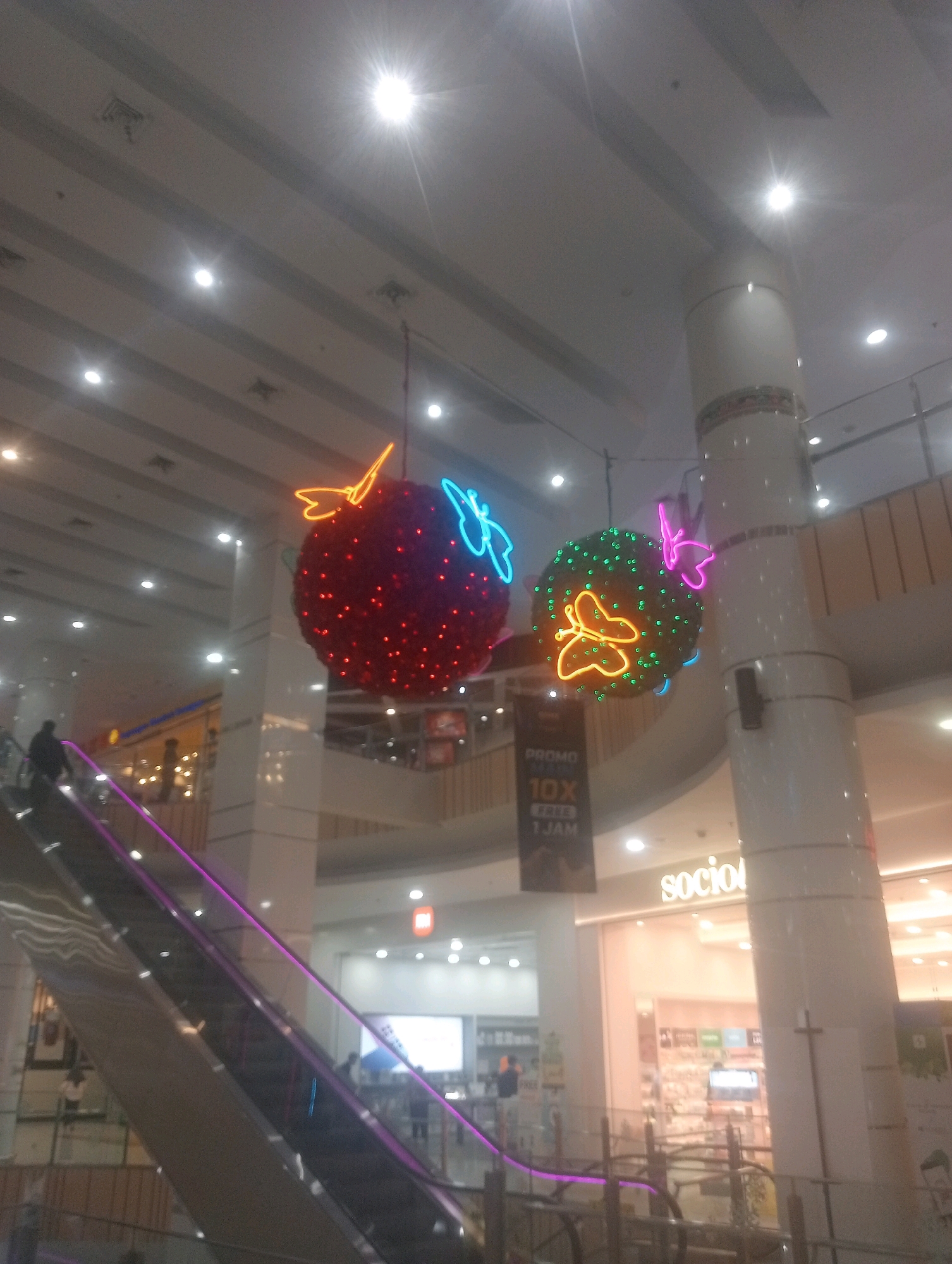 This is a photograph taken inside a shopping mall in Central Jakarta, Indonesia. The image focuses on decorative elements suspended from the ceiling, specifically two large spherical ornaments. The ornament on the left is predominantly red, adorned with numerous small red lights, and features a neon butterfly decoration in bright blue. The ornament on the right is green, similarly decorated with small lights, and has a neon butterfly in orange and another in pink. Below the ornaments, a portion of an escalator with purple lighting is visible on the left side of the frame. A few individuals are present on the escalator and in the background, appearing to be shoppers. The overall environment suggests an indoor shopping center, with the high ceiling and illuminated decor indicative of a festive or decorative display.
