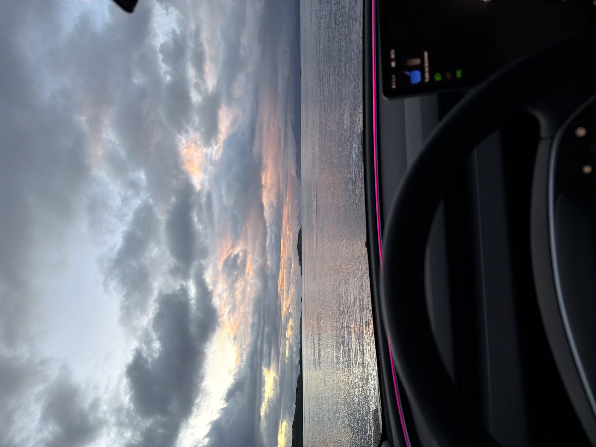 A view from inside a car, looking out at a body of water under a cloudy sky. The sky is filled with dramatic, dark gray clouds, with streaks of pink and orange light breaking through near the horizon, indicating either sunrise or sunset. The water is calm and reflects the colors of the sky. In the distance, there are islands or landmasses visible. The interior of the car shows the top of a steering wheel and a dashboard with a screen displaying some information. A pink ambient light runs along the top of the dashboard. The scene appears to be on a coastal road near Harstad, Norway, given the presence of water, islands, and the characteristic dramatic sky.