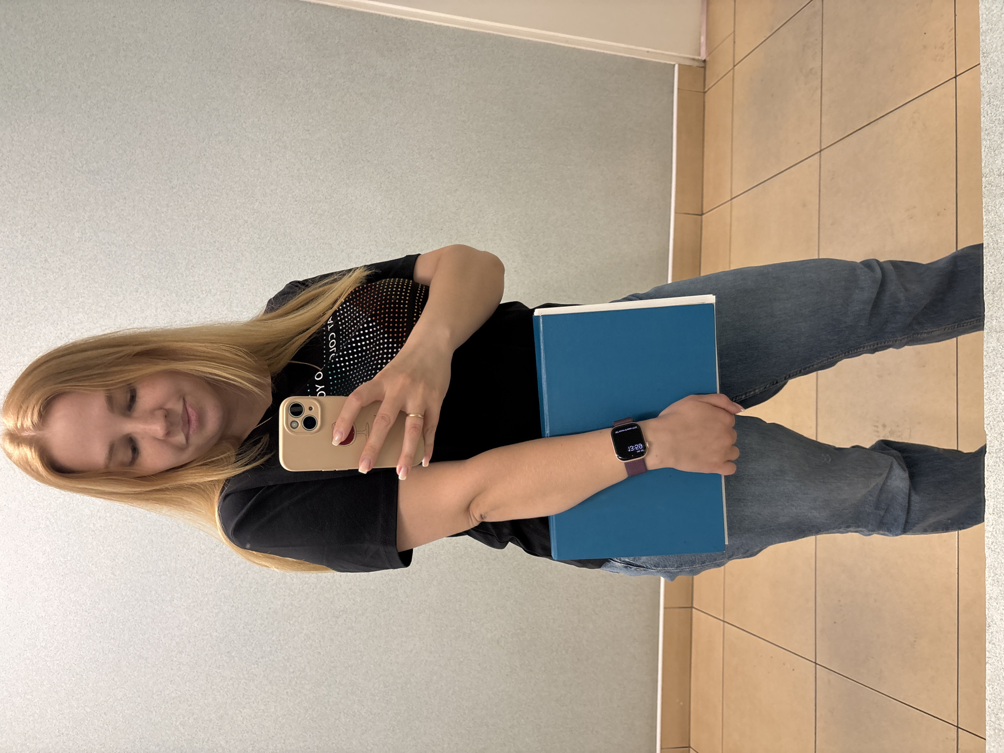 A woman stands indoors against a plain wall and tiled floor. She holds a blue book in her left hand and a tan-colored smartphone in her right. She is wearing a black t-shirt and jeans, and a smartwatch is on her left wrist. Her blond hair is long and loose. The image provides no indication of the location being Podosenikha, Russia.